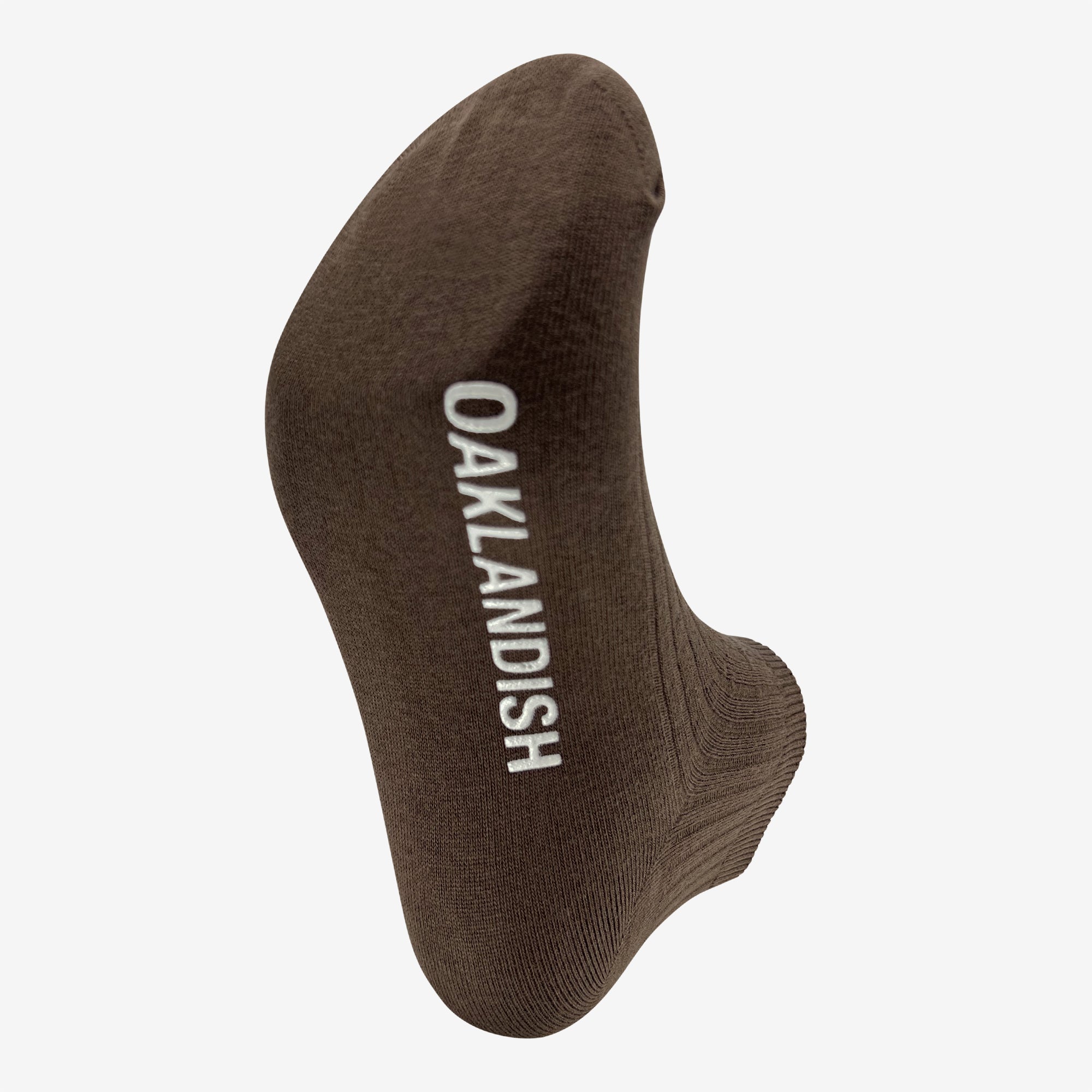 The Oaklandish Grip Low Crew Sock is a single brown cotton low crew sock with the brand name "OAKLANDISH" in white capital letters running vertically. Photographed on a white background, the image highlights the heel and toe portions of the sock, emphasizing its fabric texture and simple design.
