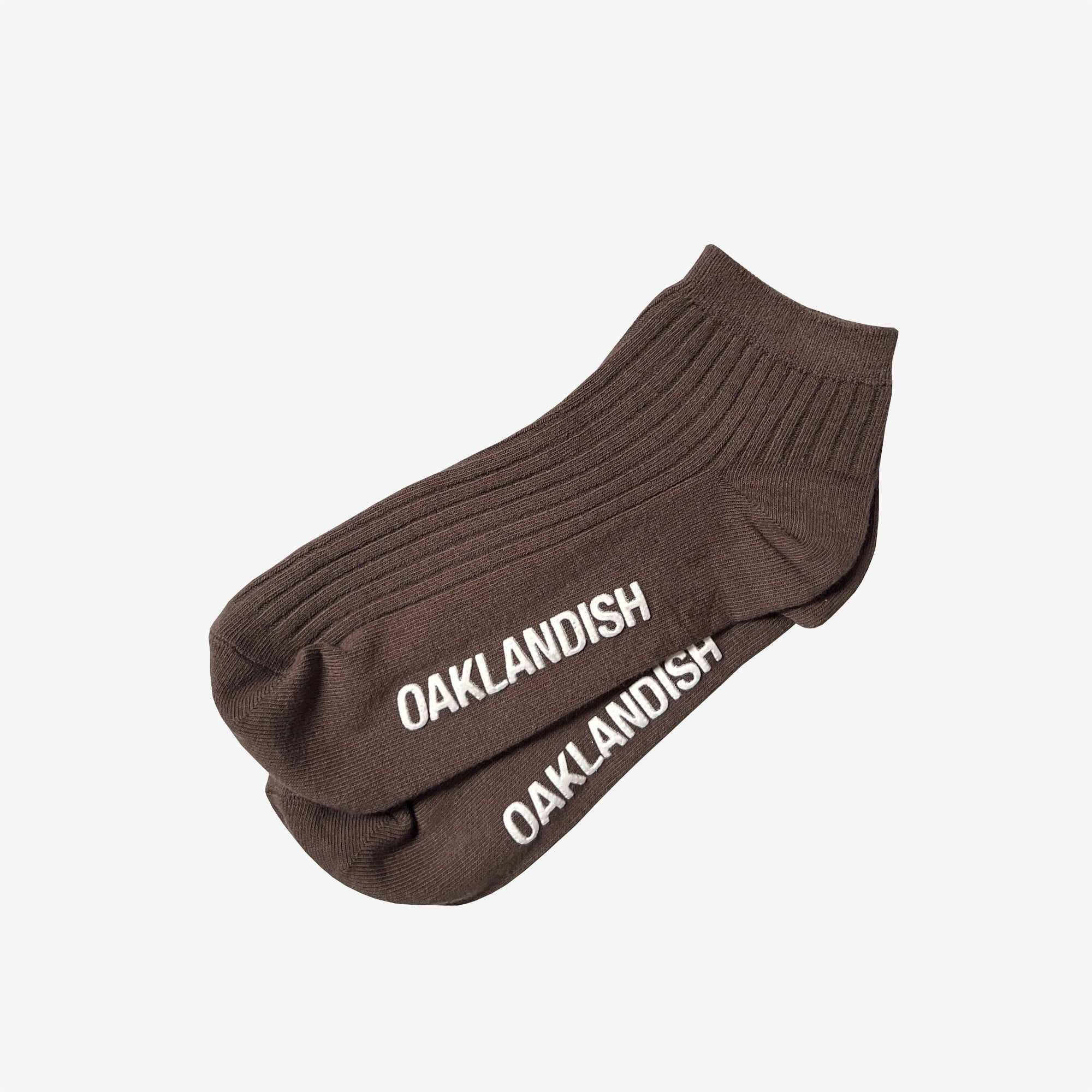 A pair of the dark brown Grip Low Crew Socks by Oaklandish, featuring ribbed fabric and white "OAKLANDISH" text embroidered near the toes. These cotton socks are neatly arranged, overlapping each other on a light gray background.