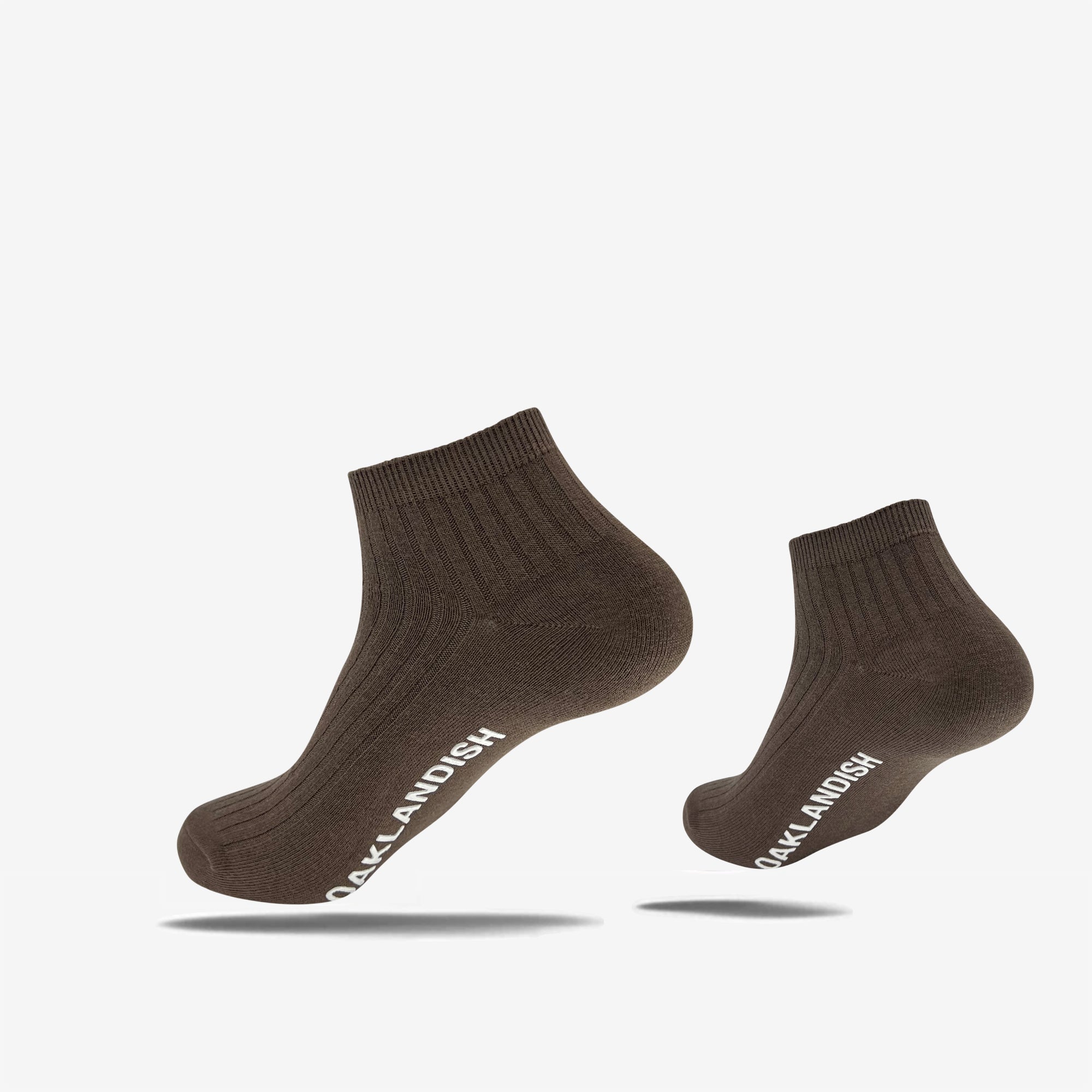 Two dark brown, cotton Grip Low Crew Socks by Oaklandish with a ribbed texture are displayed against a plain white background. The socks feature the text "OAKLANDISH" in white lettering along their sides. The image shows one sock elevated and slightly angled, while the other is positioned upright.