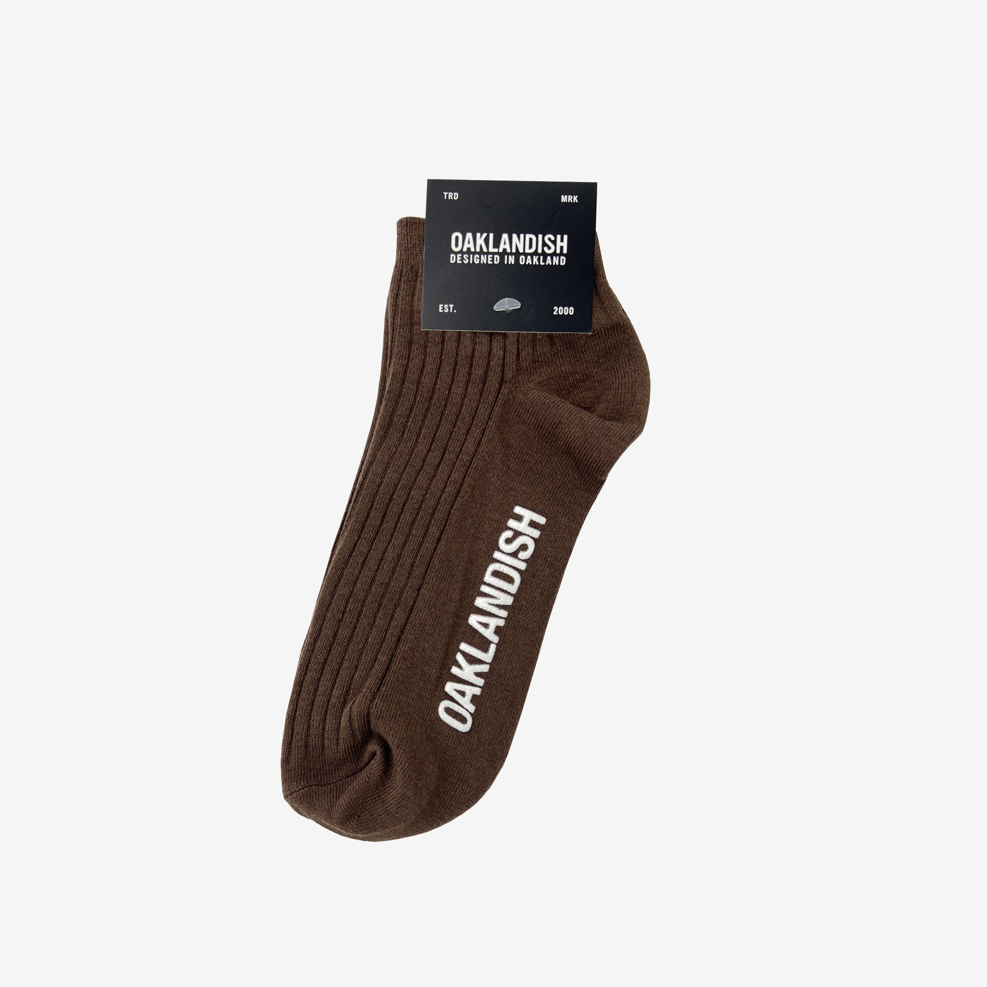 A single brown Oaklandish Grip Low Crew Sock displayed against a white background. The sock features vertical ribbing and the word "OAKLANDISH" in white capital letters along the side. Attached to the sock is a black label with white text detailing the brand name "OAKLANDISH" and other product information.