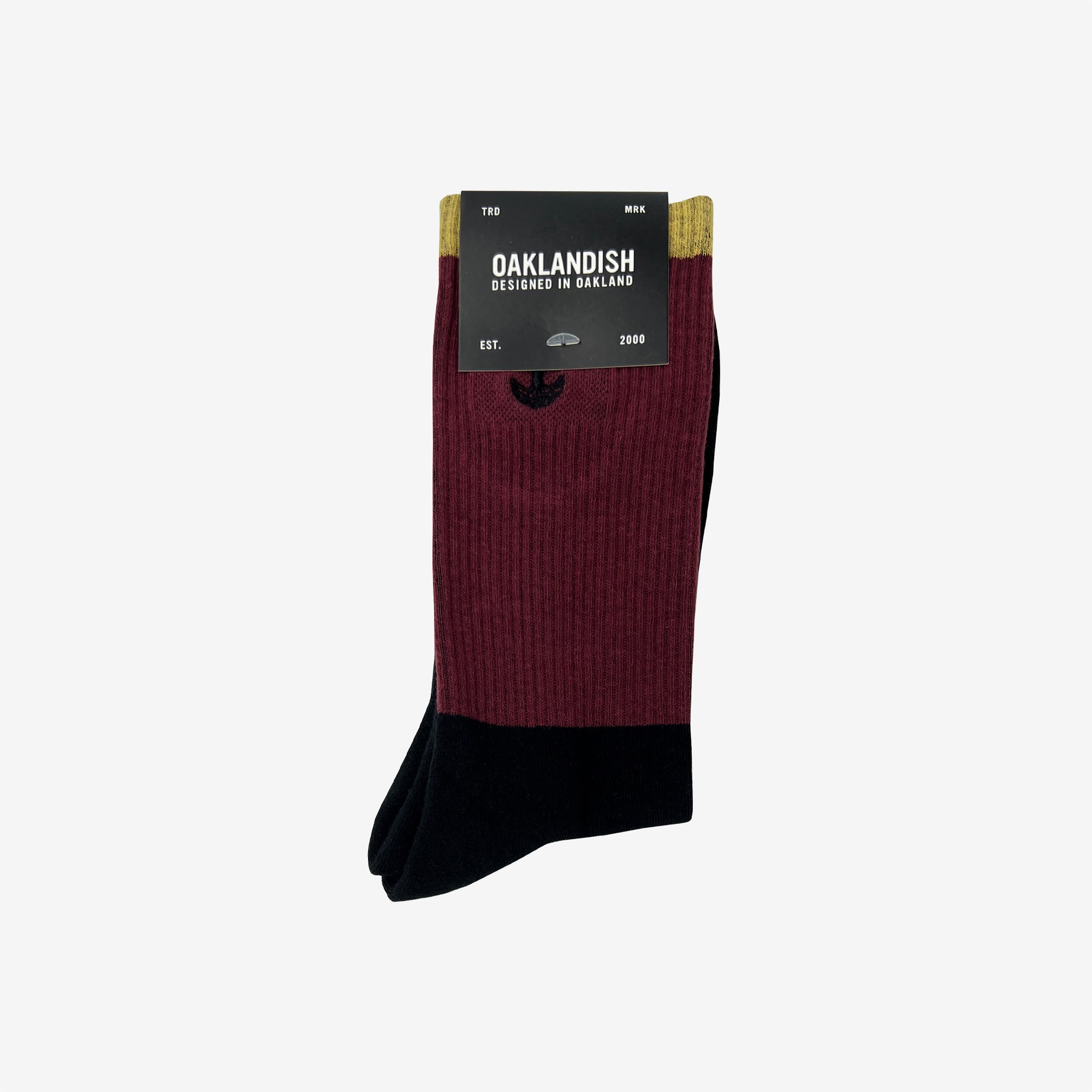 A pair of Colorblock Socks by Oaklandish features a maroon upper part and a black lower part. The packaging label reads "OAKLANDISH, Designed in Oakland, Est. 2000," and includes an embroidered Oaklandish logo along with an emblem of oak trees. Size is indicated as "TBD" and material as "MRR.