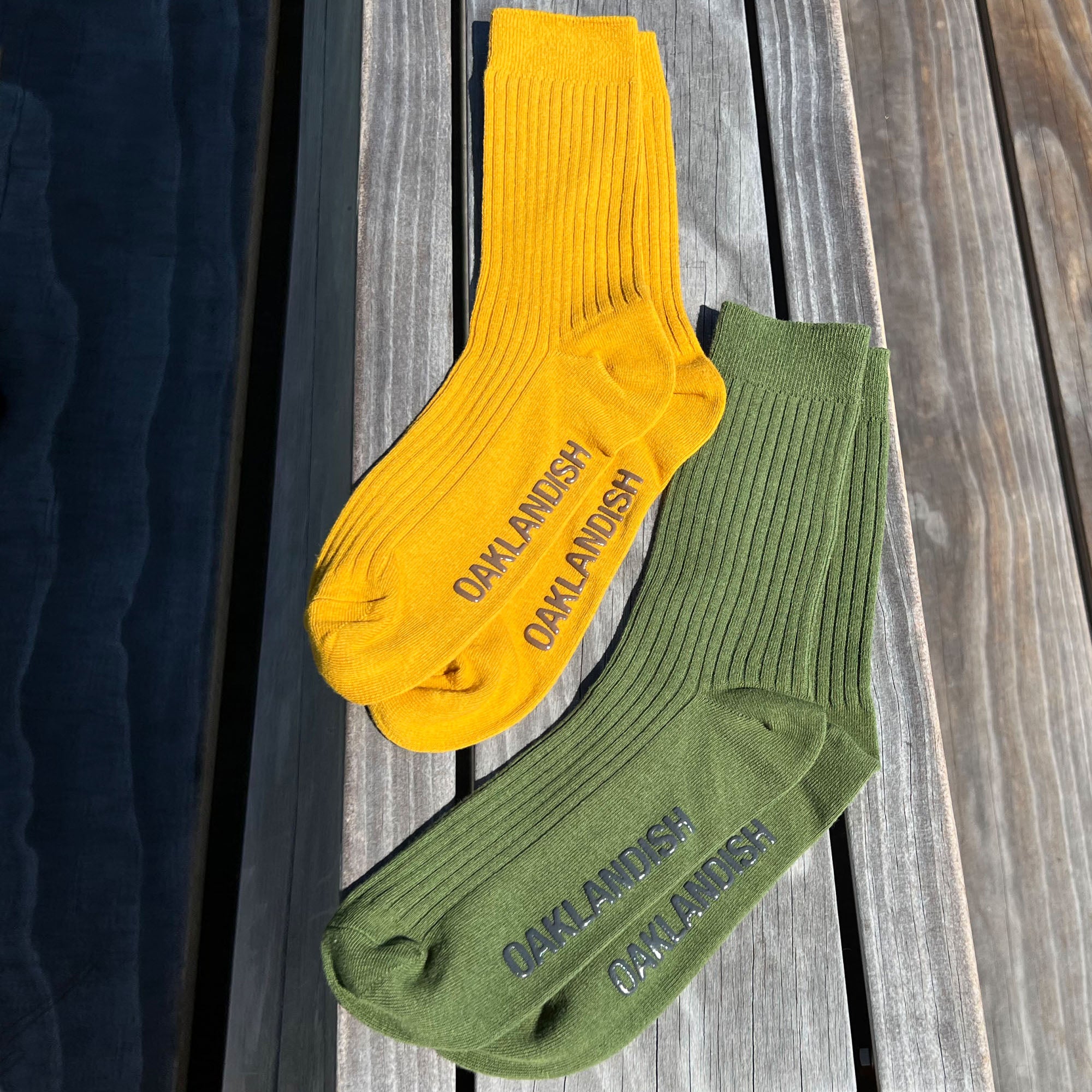 On a wooden surface lie two pairs of Oaklandish Grip Crew Socks. One pair is bright yellow, featuring the word "OAKLANDISH" printed in black near the toes, while the other pair is olive green with the same text printed in dark green. Both stylish socks come in one size that fits most.