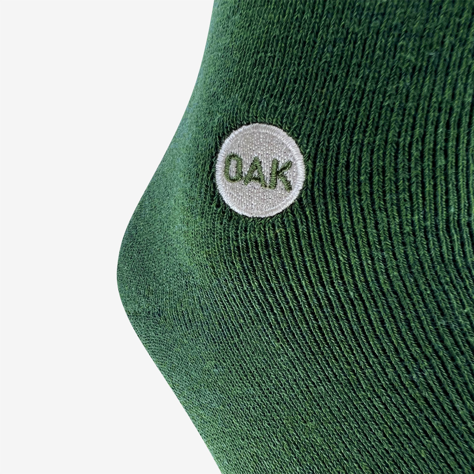 A close-up of a green fabric, likely from an Oaklandish Plush Oak Sock, with a textured, knitted pattern. There is a small white circular patch on the fabric with the uppercase letters "OAK" embroidered in green inside the patch. The background is plain white—plush socks for happy feet.