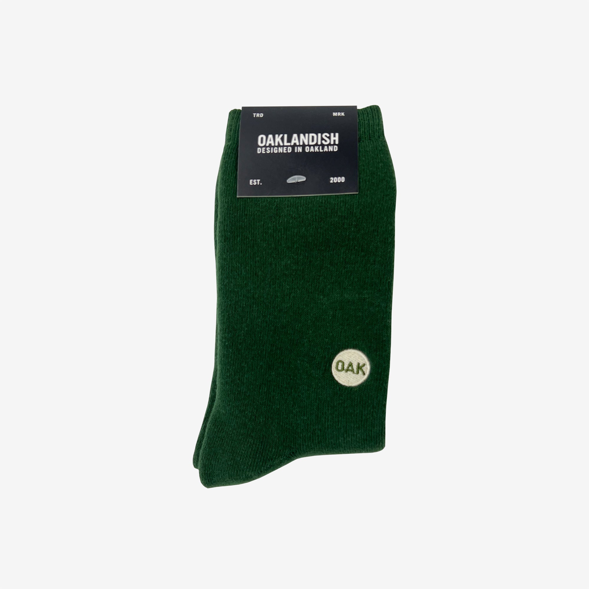 A pair of dark green Plush Oak Socks by Oaklandish are neatly folded and secured with a black label at the top. The label reads “OAKLANDISH,” “Designed in Oakland,” “Est. 2000.” These socks feature a small white circle emblem on the side, containing the letters “OAK” in green.