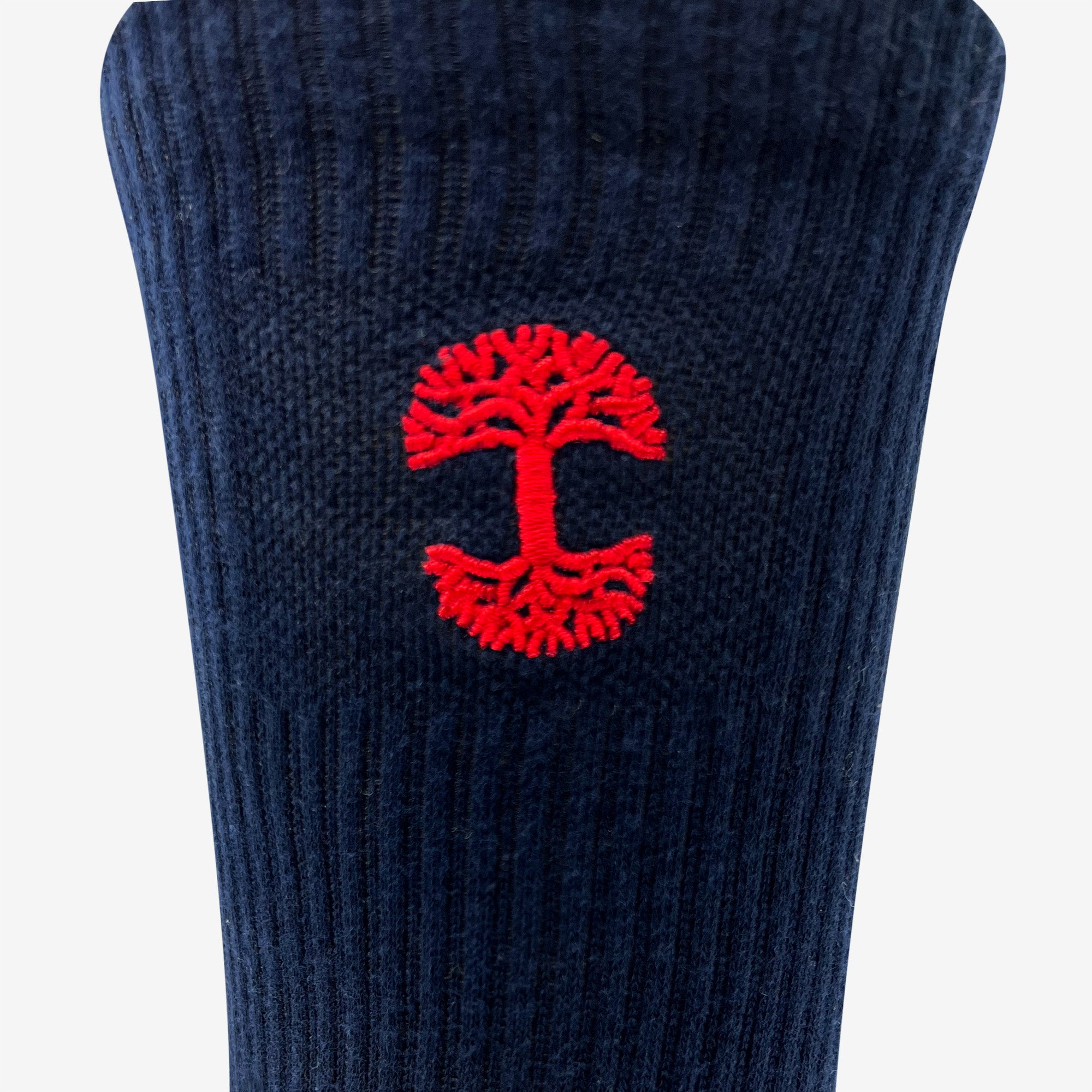 A detailed view of the Embroidered Logo Sock by Oaklandish, showcasing a dark blue ribbed knit fabric adorned with a small red embroidered Oaklandish logo—a stylized tree with mirrored branches and roots forming a circular shape—in the center. The simple background accentuates the fabric and emblem, evoking the timeless style of classic crew socks for adults.
