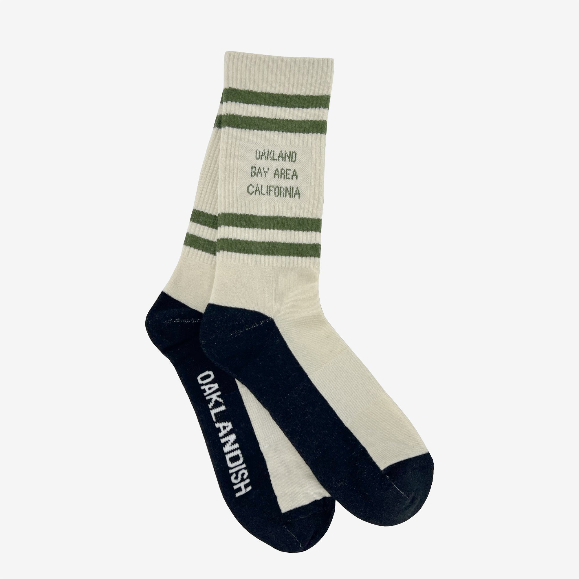 The Bay Area Stripes Sock by Oaklandish is a pair of white, cotton crew socks featuring dark green and navy blue accents. These socks have dark green stripes near the top with "Oakland Bay Area California" written below them. The toe and heel sections are navy blue, with "OAKLANDISH" written in white on the sole. Available in adult sizes.
