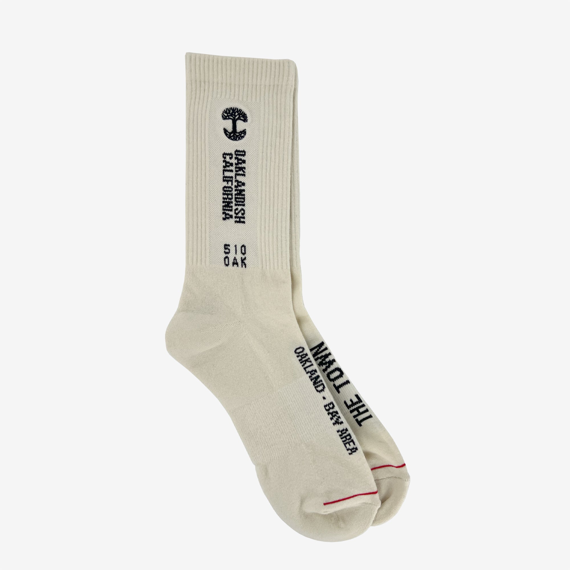 A single pair of Oaklandish Utility Socks, one size fits most, is positioned one on top of the other on a plain white background. The white crew socks feature black text that reads "THE TOWN," "OAKLAND, CALIFORNIA," "BLACK MARSH CALIFORNIA," and "510 OAK." Red stitching outlines the toes.