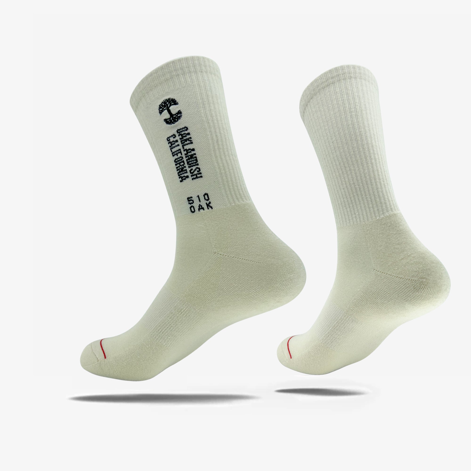 A pair of light beige utility socks from Oaklandish are shown. Each crew-length sock features a ribbed cuff and displays black text along the outer side that reads "OAK KNITISH CALIFORNIA" vertically and "510 OAK" horizontally. These Oaklandish Utility Socks, made from a cotton polyester spandex blend, are displayed upright with subtle shadows beneath them against a white background.