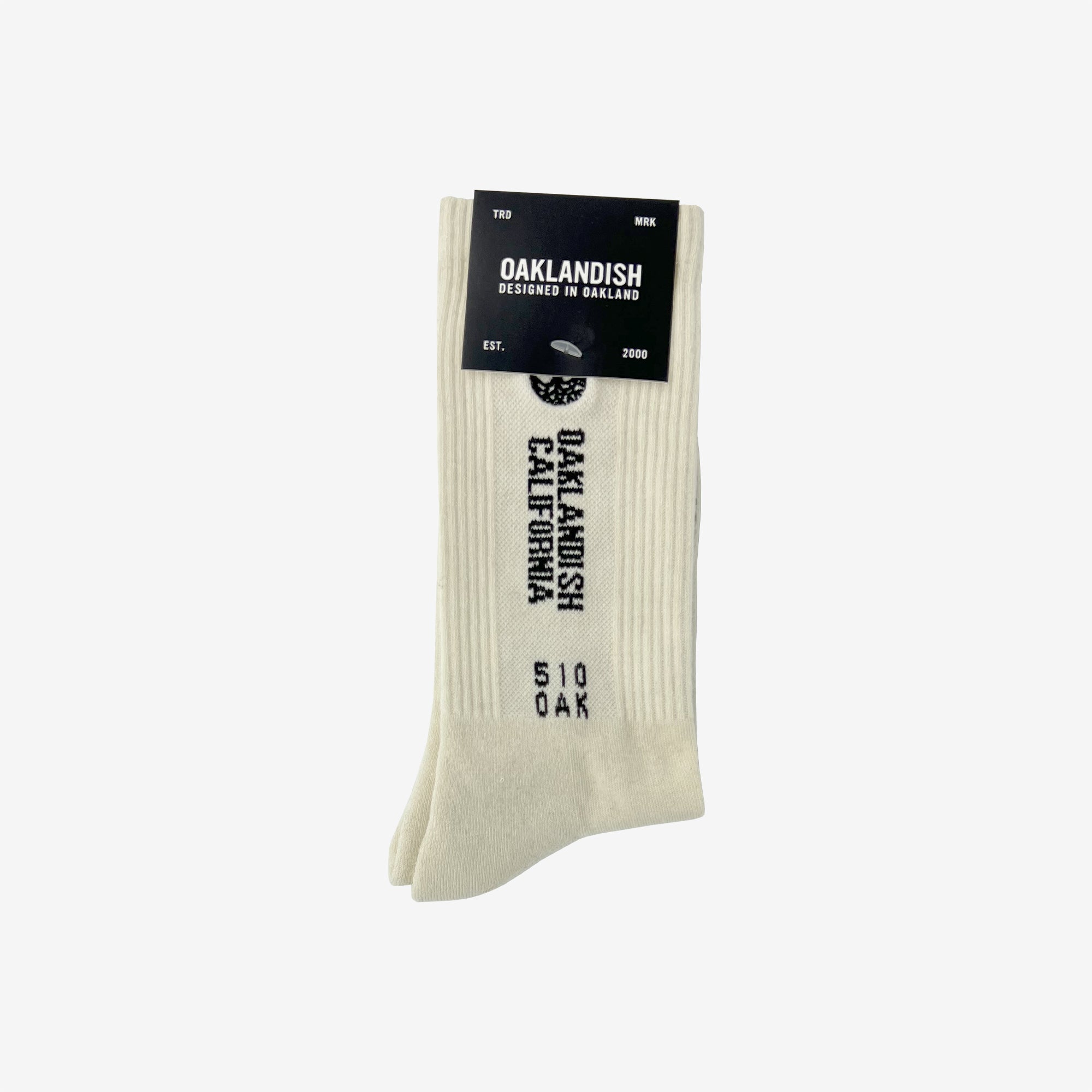 A pair of Oaklandish Utility Socks in beige, featuring the Oaklandish logo and black vertically displayed text that reads "Oaklandish California 510 OAK." The packaging at the top states "OAKLANDISH Designed in Oakland" with a tree logo, and below it reads "EST. 2000." These medium-sized socks are made from a cotton-polyester-spandex blend.