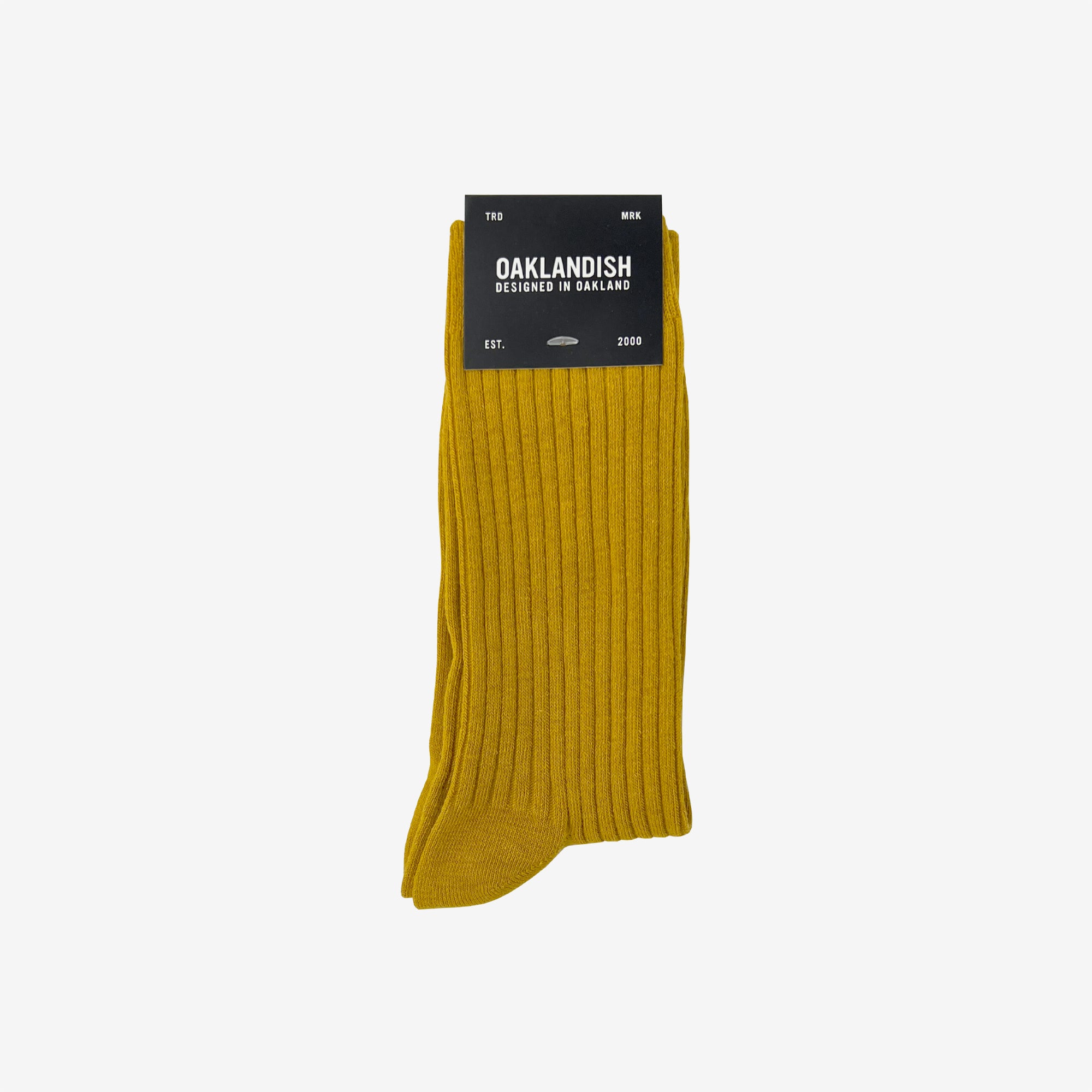 A single pair of mustard yellow Grip Crew Socks from Oaklandish is displayed against a white background. The packaging at the top prominently features the "Oaklandish" brand name in bold white text, along with additional details indicating that these cotton polyester spandex socks are designed in Oakland and available in size M/L.