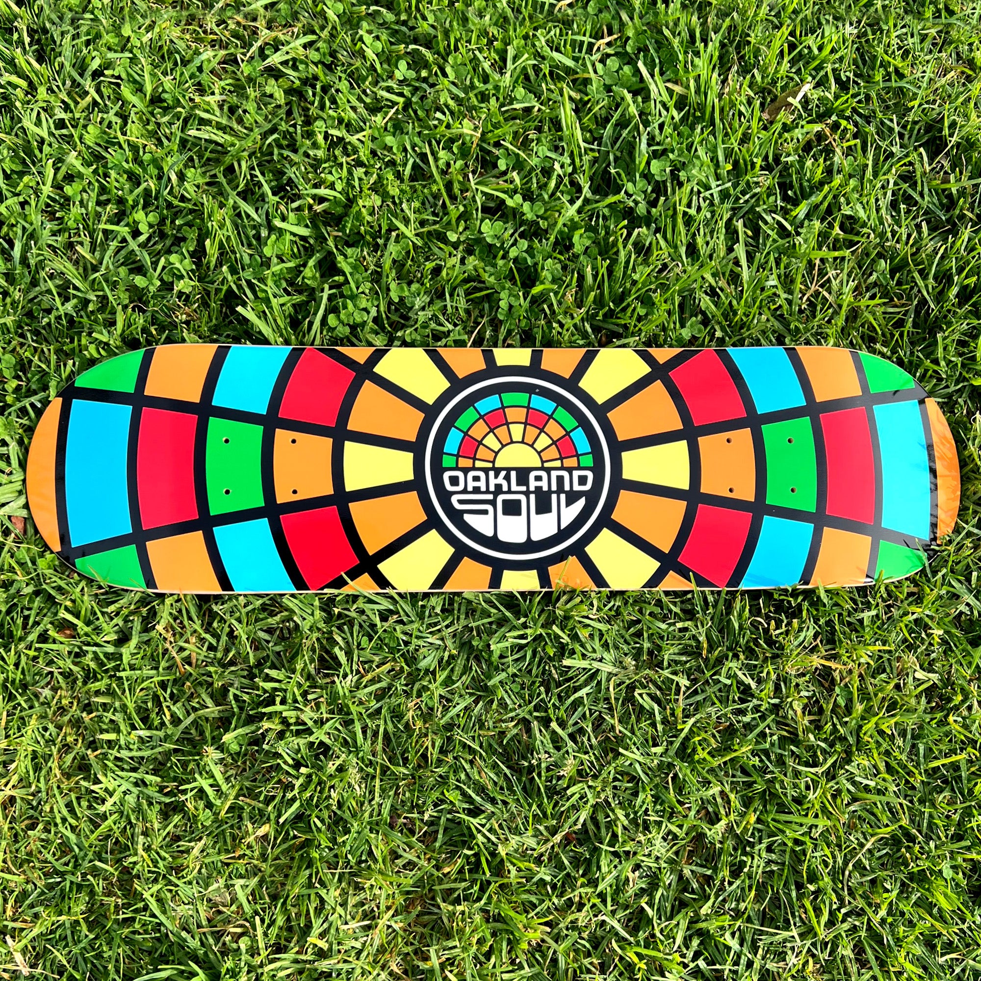 The Oakland Soul Logo Skate Deck, resting on the grass, showcases a vibrant geometric design. Constructed from durable 7-ply maple wood, it features a multicolored pattern in shades of red, orange, yellow, green, blue, and black arranged in a striking radial mosaic style. At the center is the Oakland Soul SC logo with “OAKLAND” in white above graffiti-style letters spelling “SOUL.”