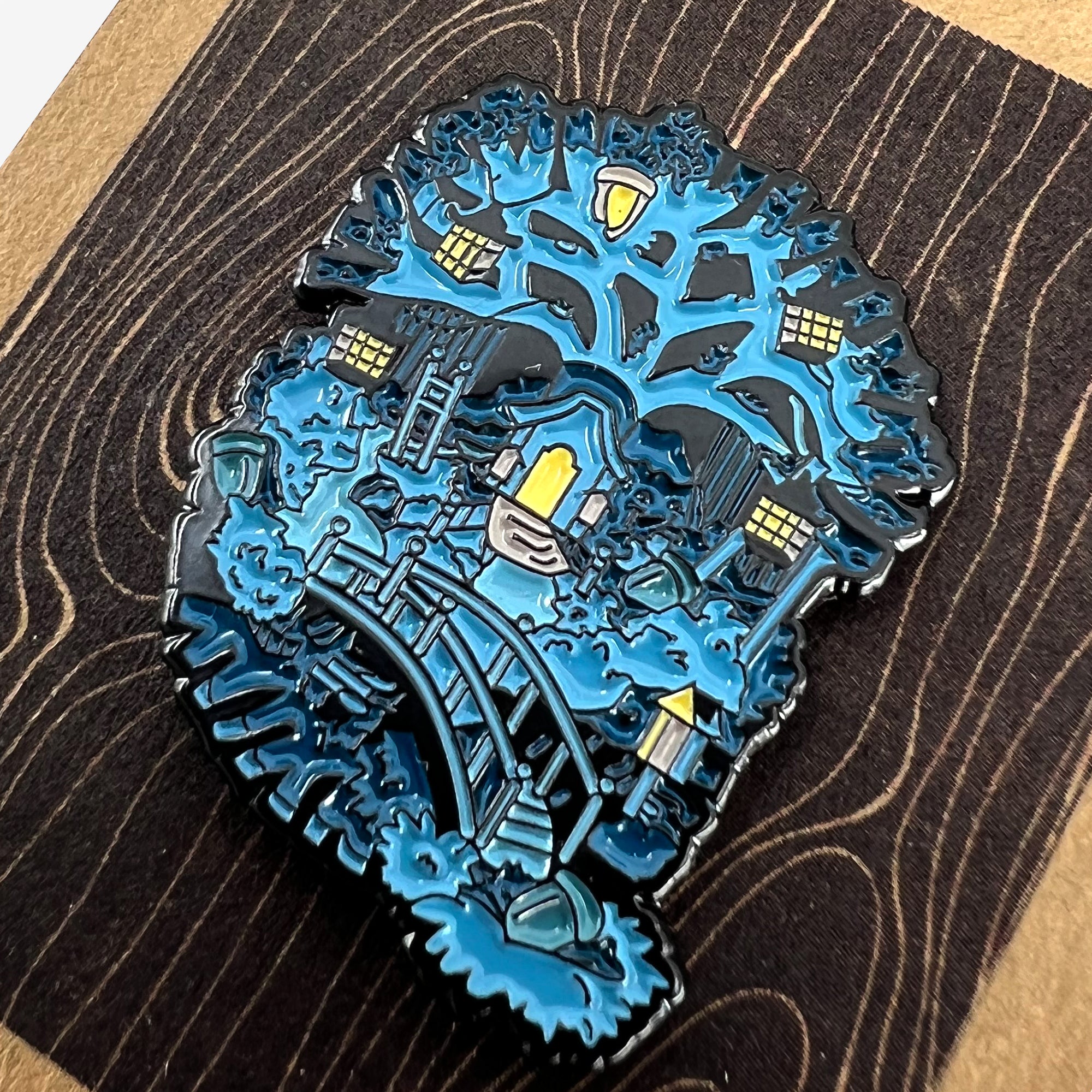 An intricate Enamel Pin - Treehouse by Oaklandish depicts a serene night scene with a treehouse. The tree in this Oakland-inspired design is adorned with lantern-like windows, and below it, a wooden bridge spans across a small creek. This accessory features shades of blue, capturing a calm, whimsical atmosphere.