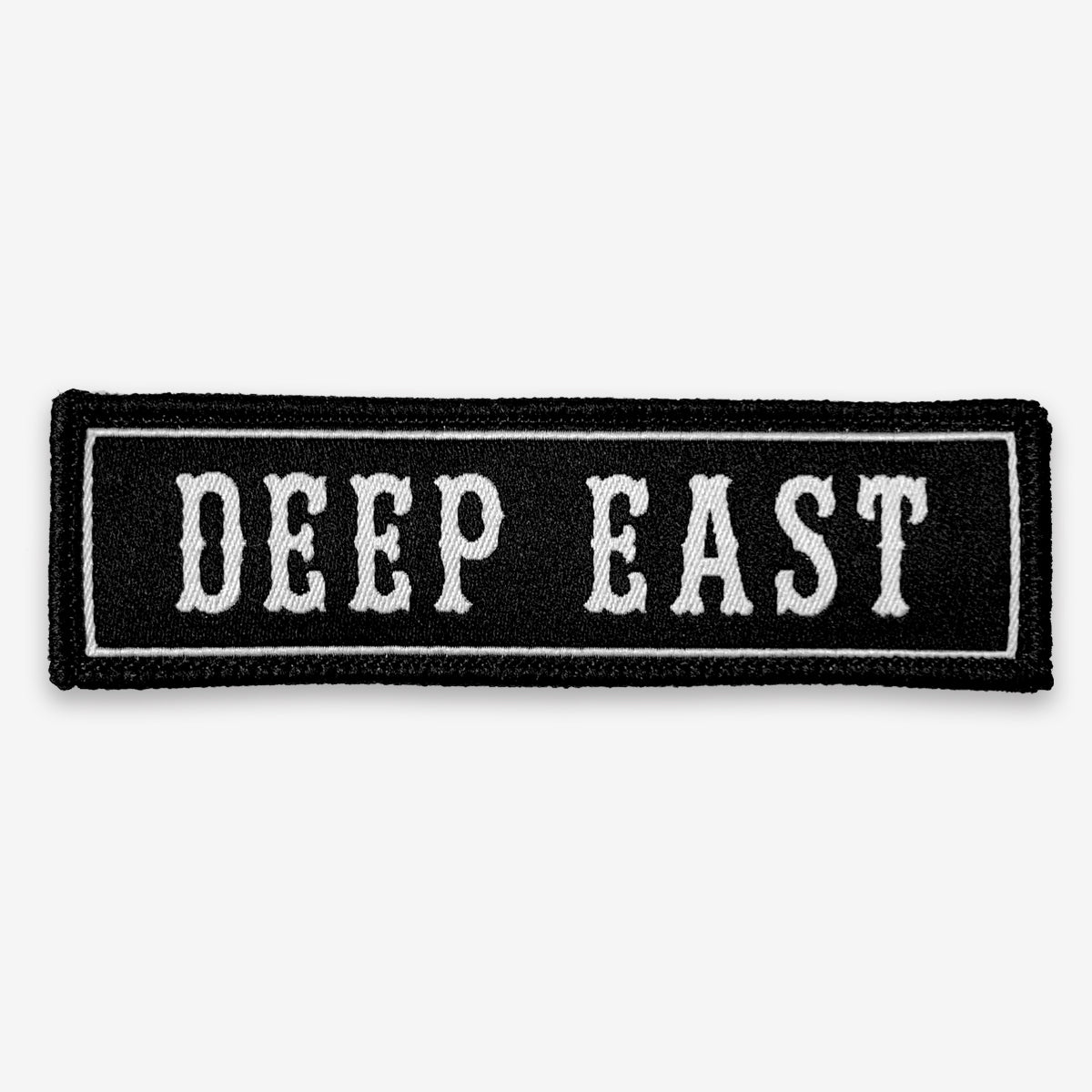 The "Deep East Patch" by Oaklandish is a black, rectangular woven iron-on patch featuring "DEEP EAST" embroidered in white capital letters, outlined by a white rectangular border with black edging. Ideal for fans of Oakland's vibrant spirit, this accessory stands out against the overall white background.