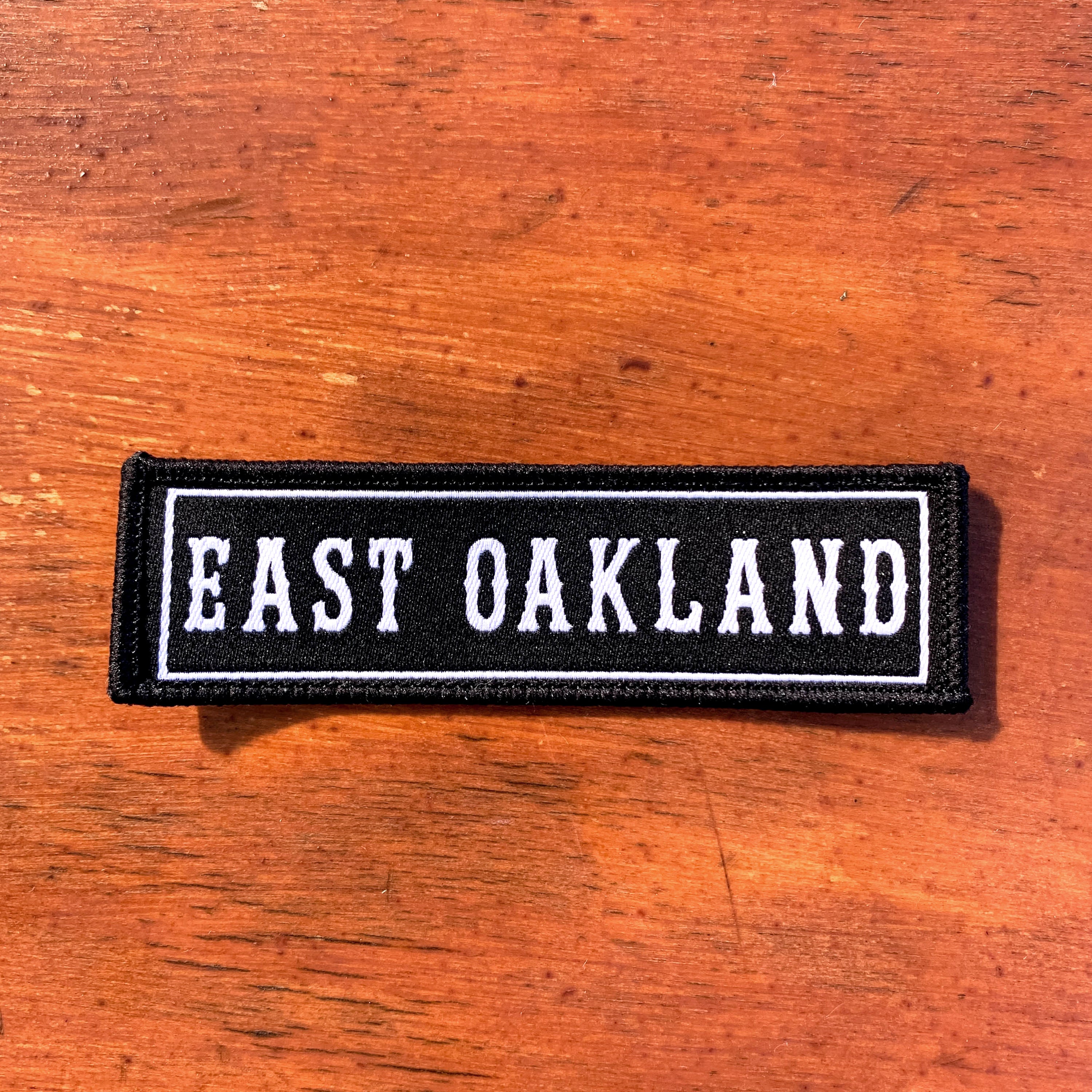 A rectangular East Oakland Patch by Oaklandish, featuring a black background and a white border, lays on a wooden surface. The patch proudly displays the words "EAST OAKLAND" in uppercase, white, bold letters. The wood beneath it showcases a natural grain and warm brown tones.