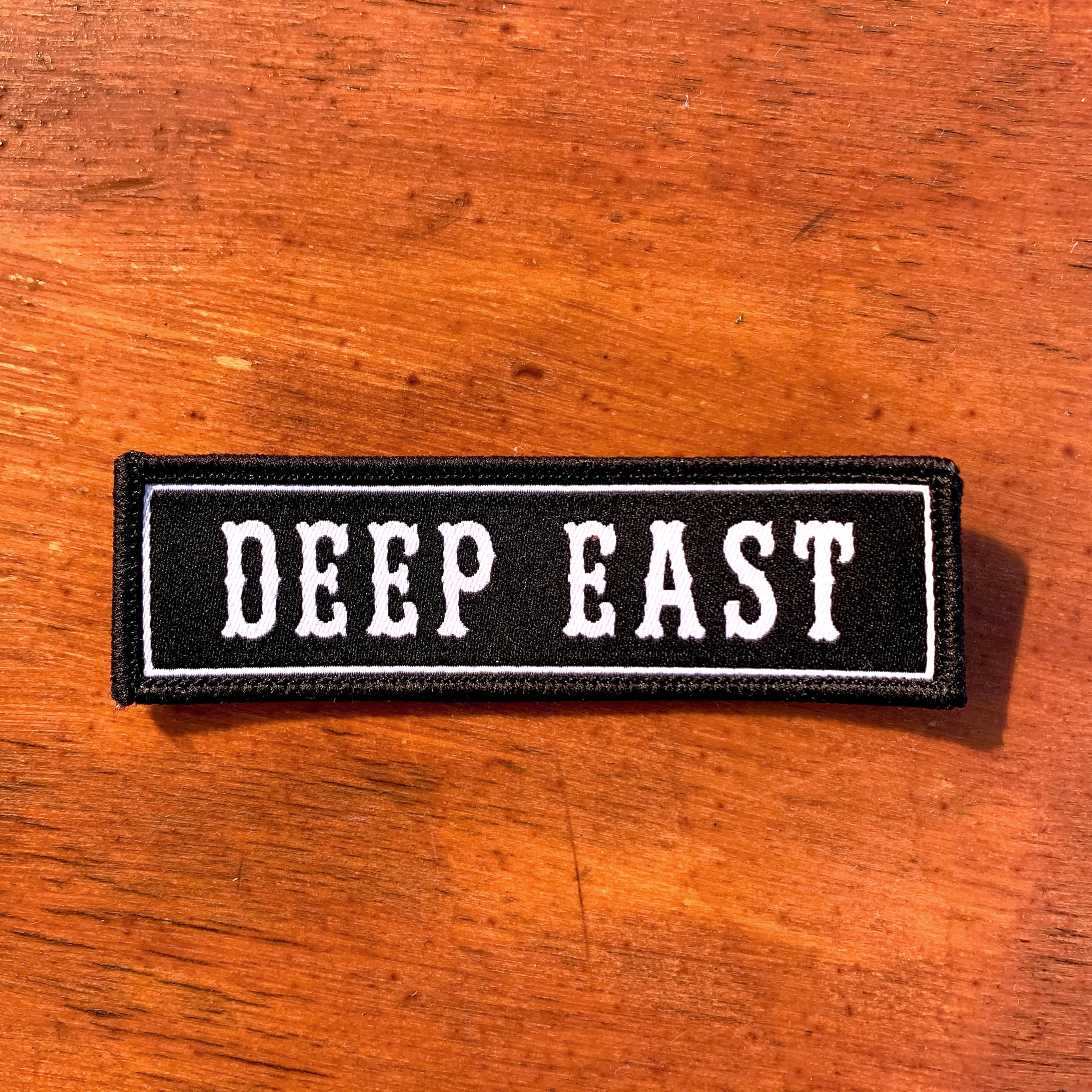 Introducing the Deep East Patch by Oaklandish: a rectangular black woven iron-on patch featuring the words "DEEP EAST" embroidered in bold, white block font and framed by a white border. This stylish accessory is perfect for proudly representing the spirit of Oakland and is showcased on a wooden surface.