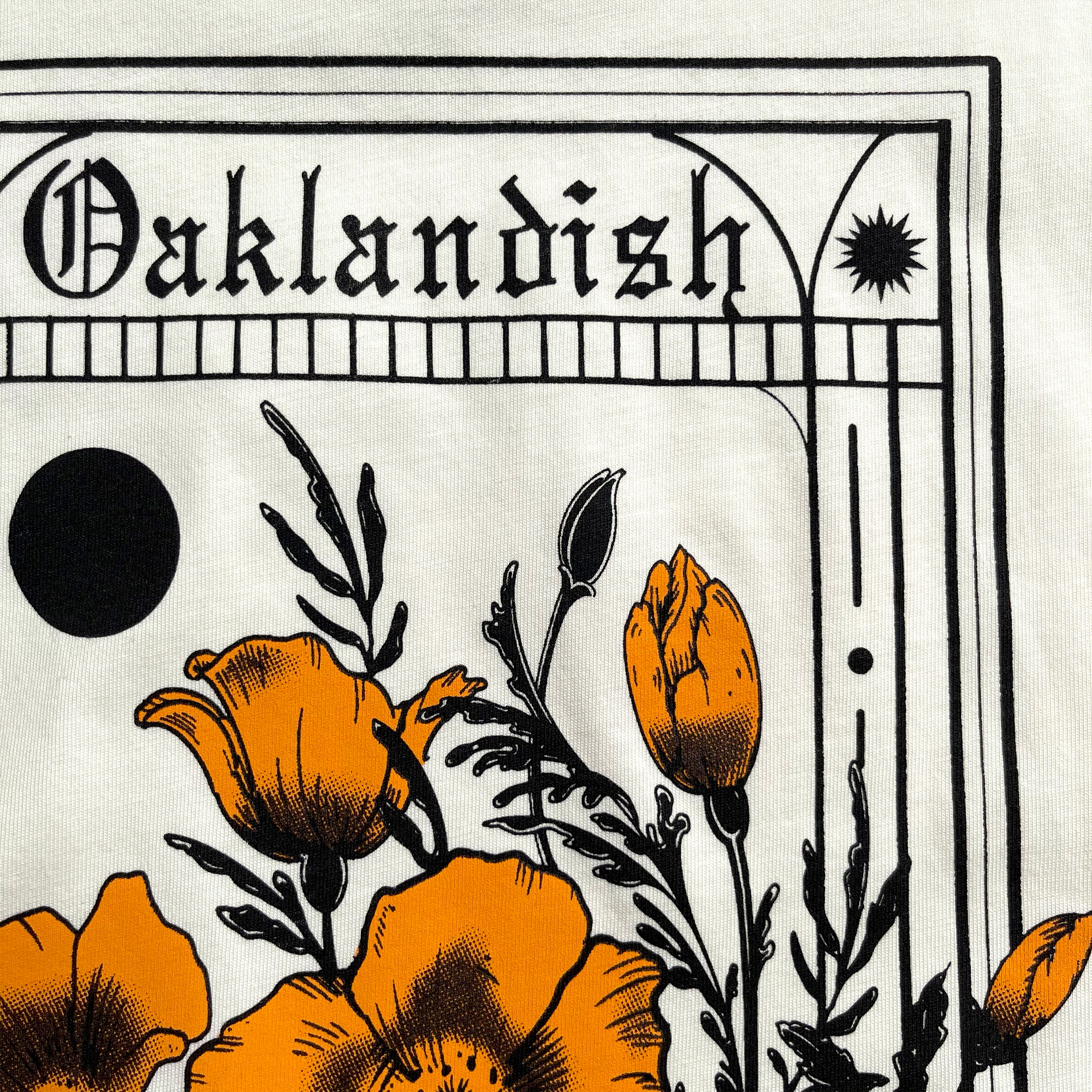 The Women's Blossom Tee from Oaklandish features an intricate black and white illustration with bold orange poppies and foliage on its cream-colored front. Above the flowers, "Oaklandish" is displayed in a Gothic font, framed by geometric shapes and lines, including a large black circle and a small black star. This t-shirt is made of 100% cotton.