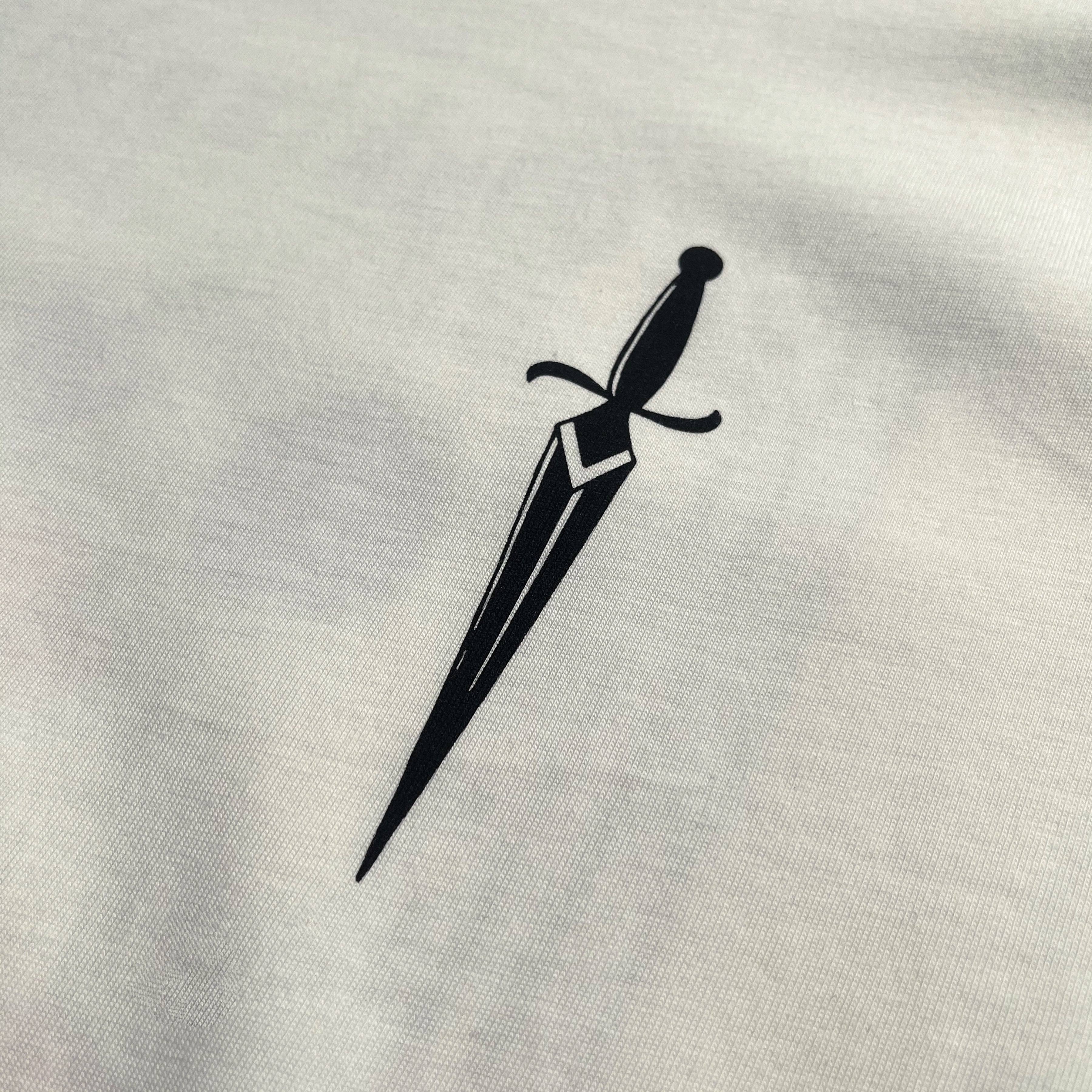 The Blossom Tee by Oaklandish features a minimalist black dagger design centered on a plain white, 100% cotton background. The dagger showcases a straight blade with a geometric pattern near the hilt and a simple crossguard, highlighting the clean, bold contrast on this classic fit t-shirt.