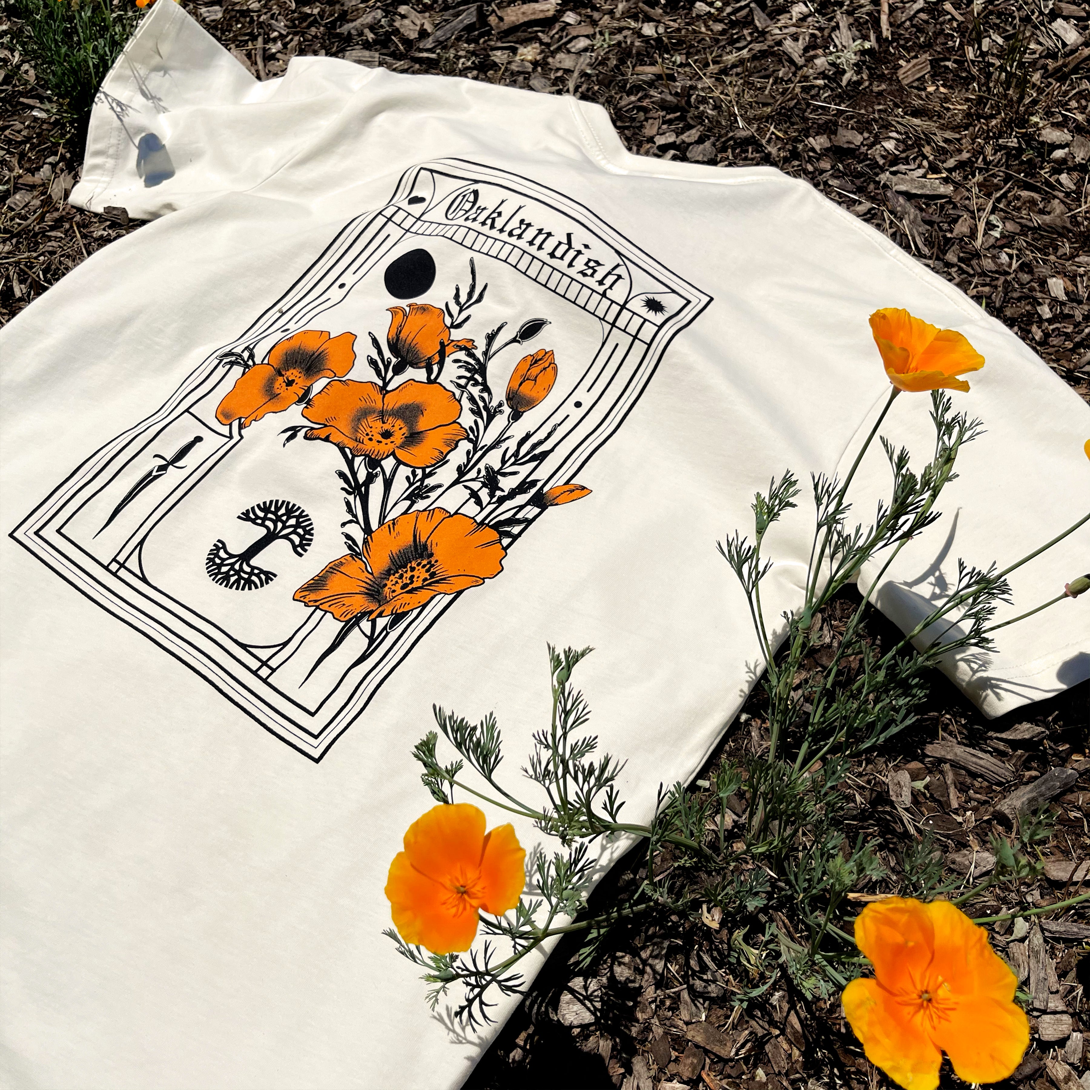 The Blossom Tee by Oaklandish is a white, 100% cotton classic fit t-shirt with an artistic design featuring vibrant orange poppies and a caterpillar framed by intricate black lines. The shirt lies on soil scattered with mulch, surrounded by real orange California poppies that complement its floral design. The top of the design showcases the brand name "Oaklandish.