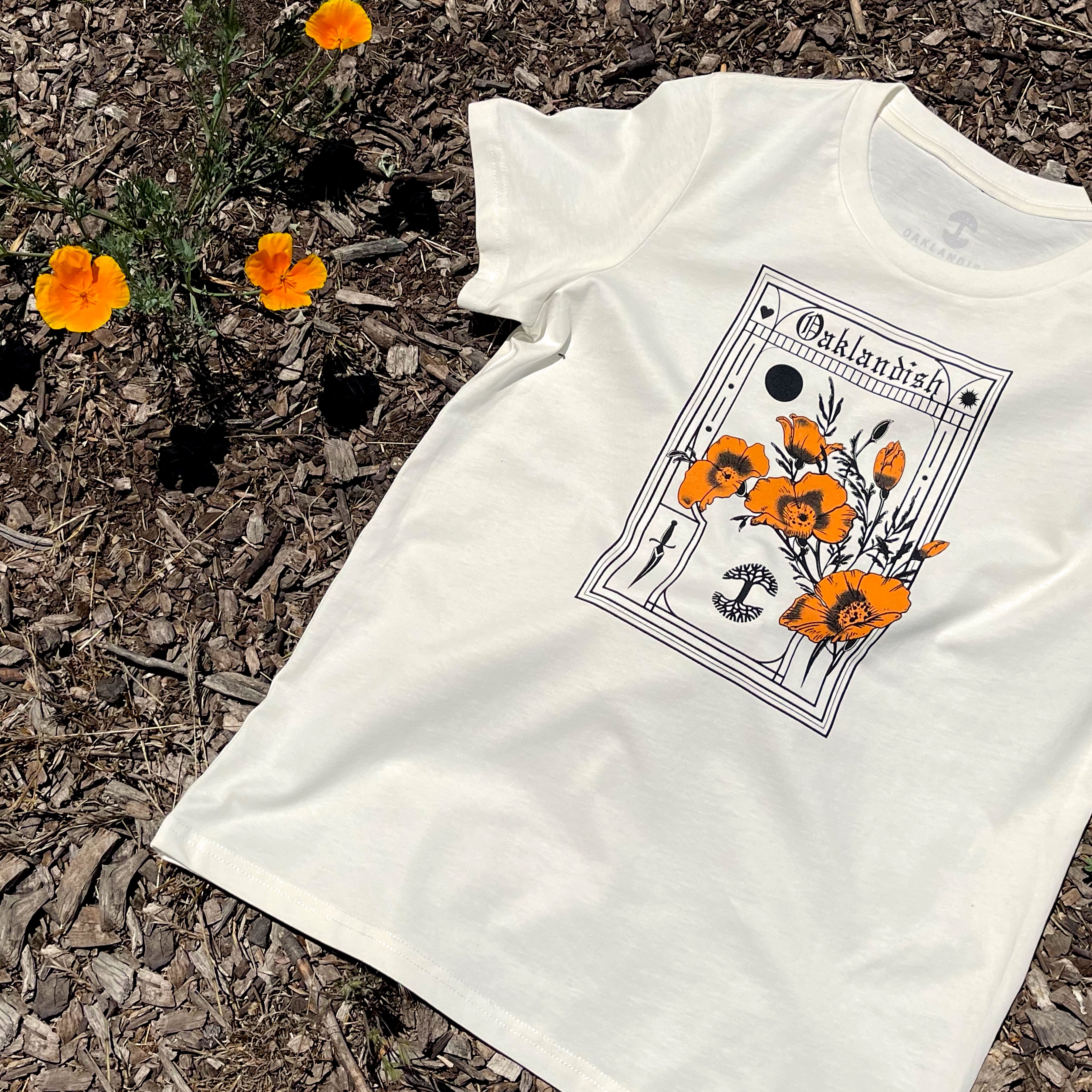 The Women's Blossom Tee by Oaklandish, made from 100% cotton in a classic fit, rests on wood chips surrounded by blooming orange flowers. The white T-shirt features an intricate botanical illustration of poppies and the word "Oaklandish" at the top, standing out against the vibrant floral background.