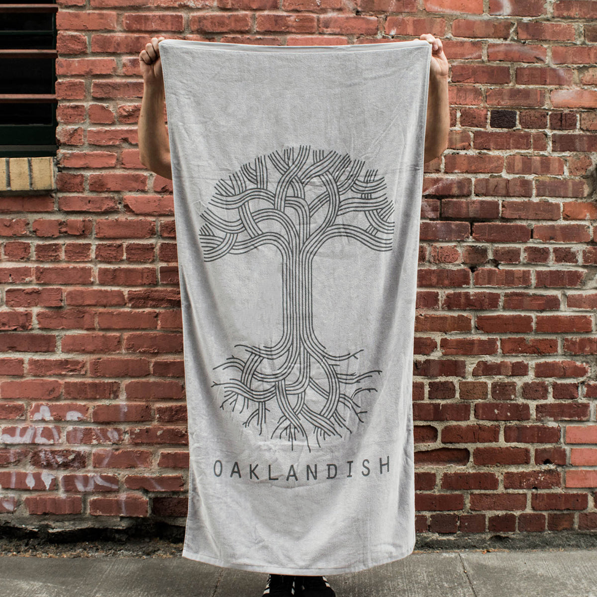 A person is holding up the Oaklandish Classic Logo Beach Towel, an oversized, super soft towel in light grey. It features a stylized black tree design with intertwined branches forming an intricate pattern and mirrored roots. Below the tree, "OAKLANDISH" is printed in black. Behind the person is a red brick wall.
