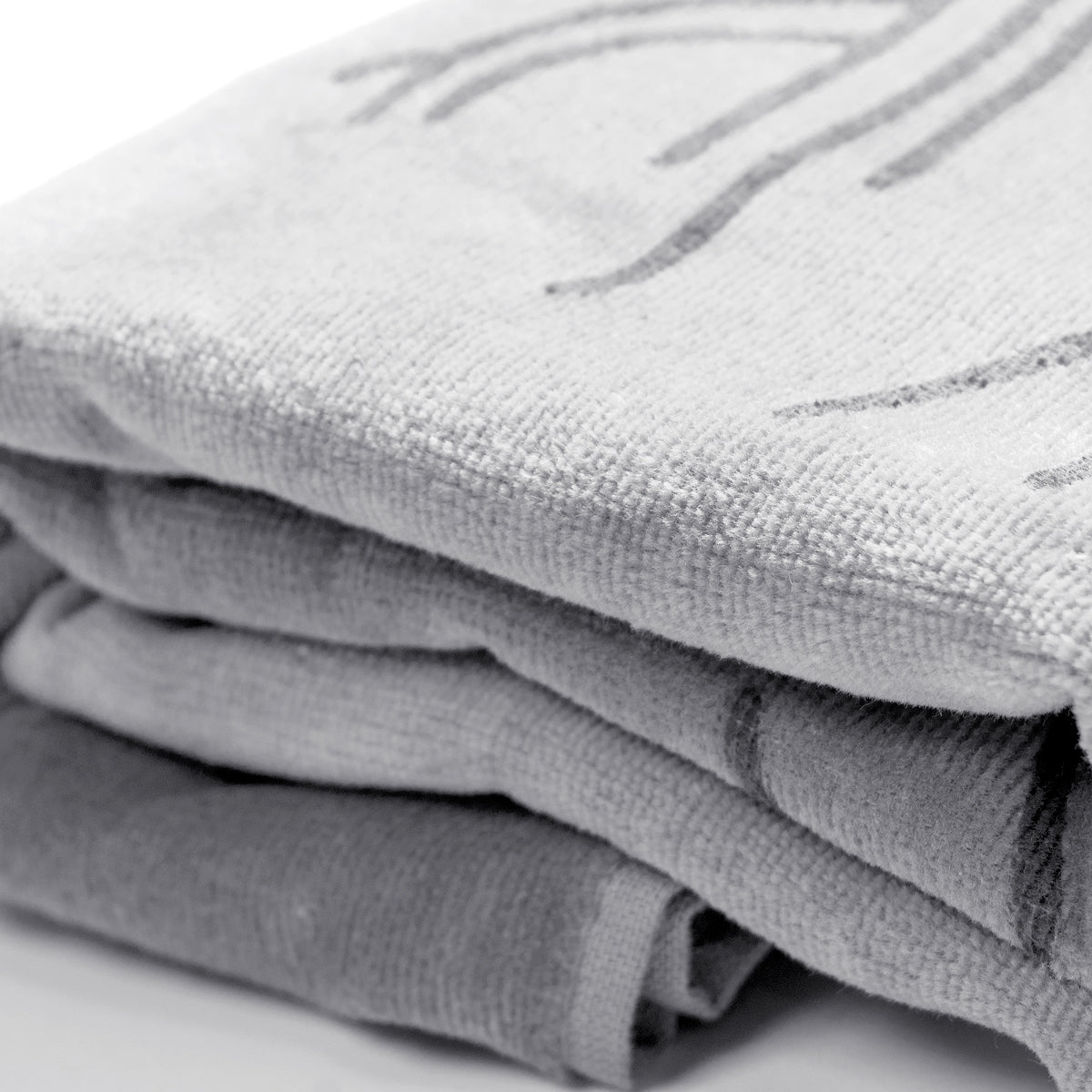 A neatly arranged stack of three folded Classic Logo Beach Towels in various shades of gray rests on a smooth, white surface. Made from 100% ringspun cotton, these Oaklandish towels feature a textured surface with faint abstract patterns. The top towel showcases a subtle curved design, while the others are more plain and simple.