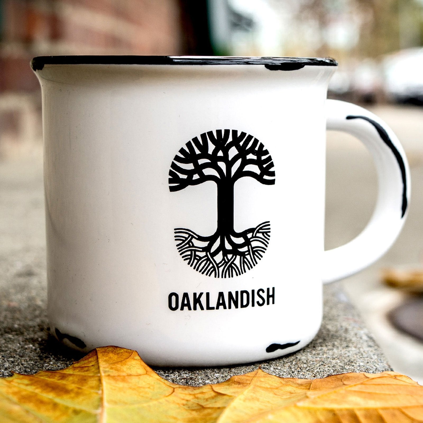 A white ceramic 15 oz Ventura Mug from Oaklandish with black rim accents sits on a concrete surface. The mug showcases a black graphic of a tree with roots extending below, with the word "OAKLANDISH" printed beneath it, symbolizing pride in the city of Oakland. Two autumn leaves are partially visible in the foreground, while the background remains blurred.