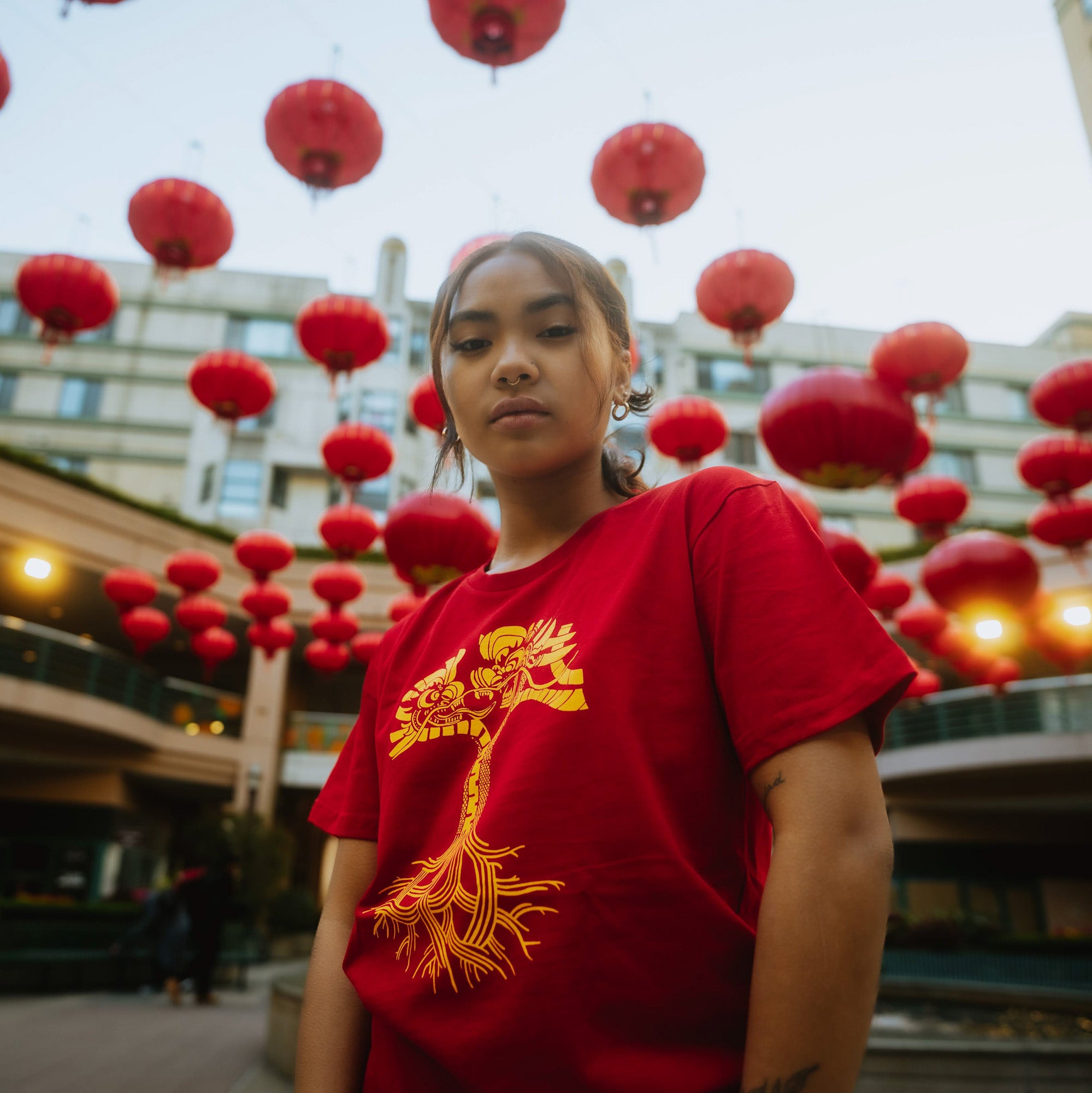 A person donning the Dragon Power Tee from Oaklandish, featuring a captivating yellow tree design on a vibrant red shirt, stands in the outdoor courtyard of Oakland Chinatown, which is beautifully decorated with numerous hanging red lanterns. The background showcases a multi-story building with balconies and windows. The person, facing the camera with a calm expression, epitomizes the rich Chinese American heritage of the area.