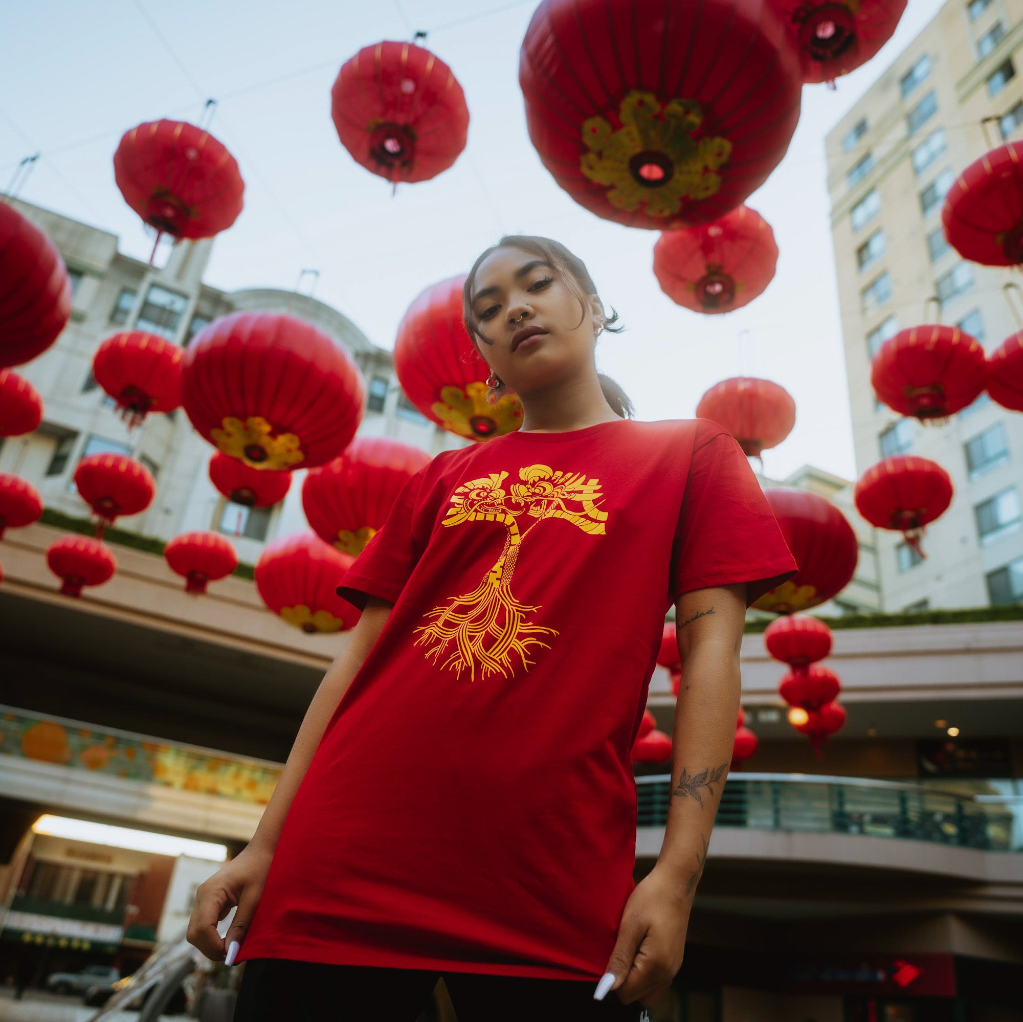 A person wearing the Dragon Power Tee from Oaklandish, featuring a striking yellow dragon design on a red shirt, stands confidently under numerous red lanterns hanging in the outdoor plaza of Oakland Chinatown. The person, celebrating their Chinese American heritage, has long hair tied back and an intricate tattoo on their forearm. Buildings and a clear sky surround the festive scene.