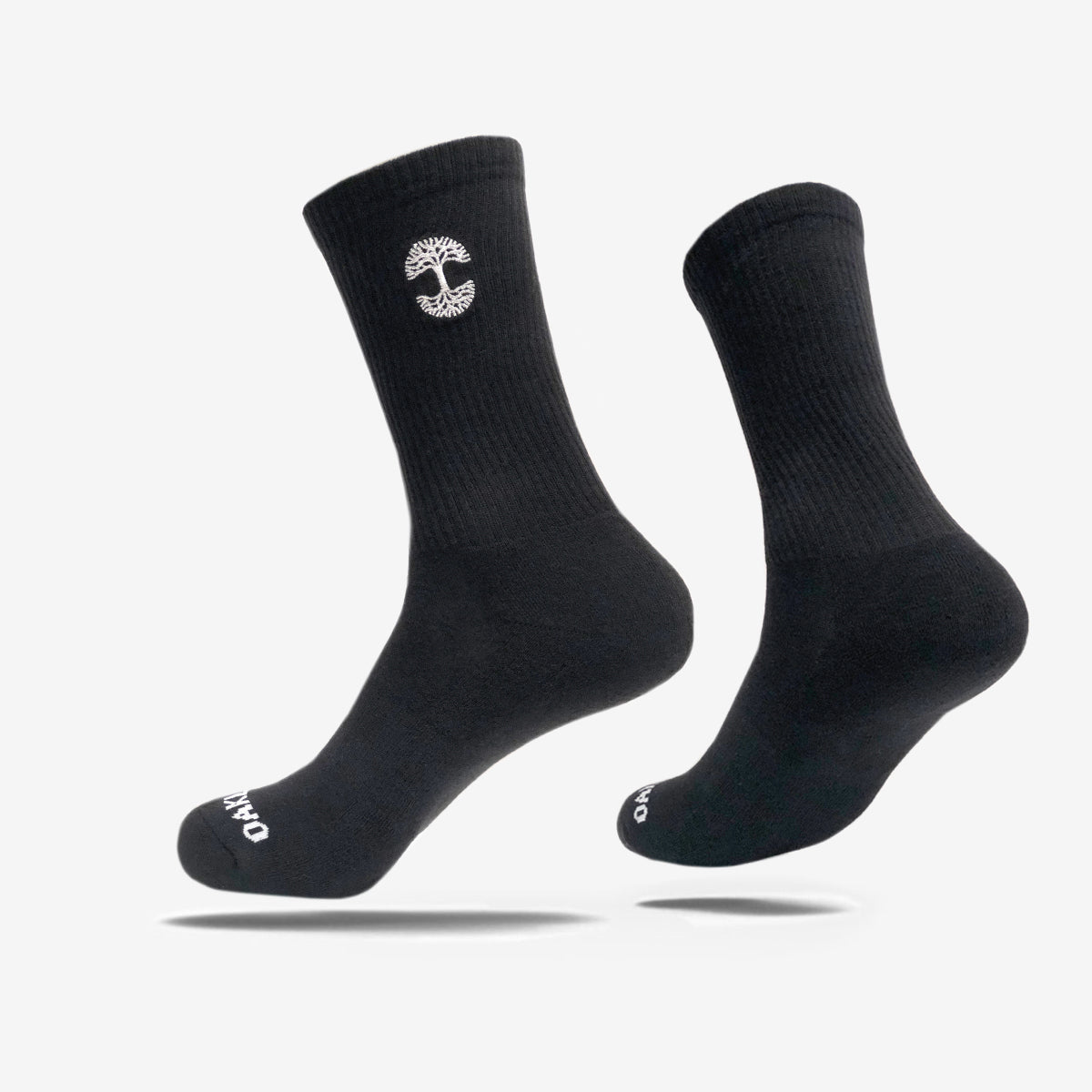 A pair of black crew socks, with one sock slightly elevated and angled to the side. The Embroidered Logo Socks feature a small, white, vertical emblem resembling a tree on the upper part and white text "OAKPO" near the toes. These Oaklandish socks come in adult sizes, boasting a ribbed texture on top and smooth finish below.