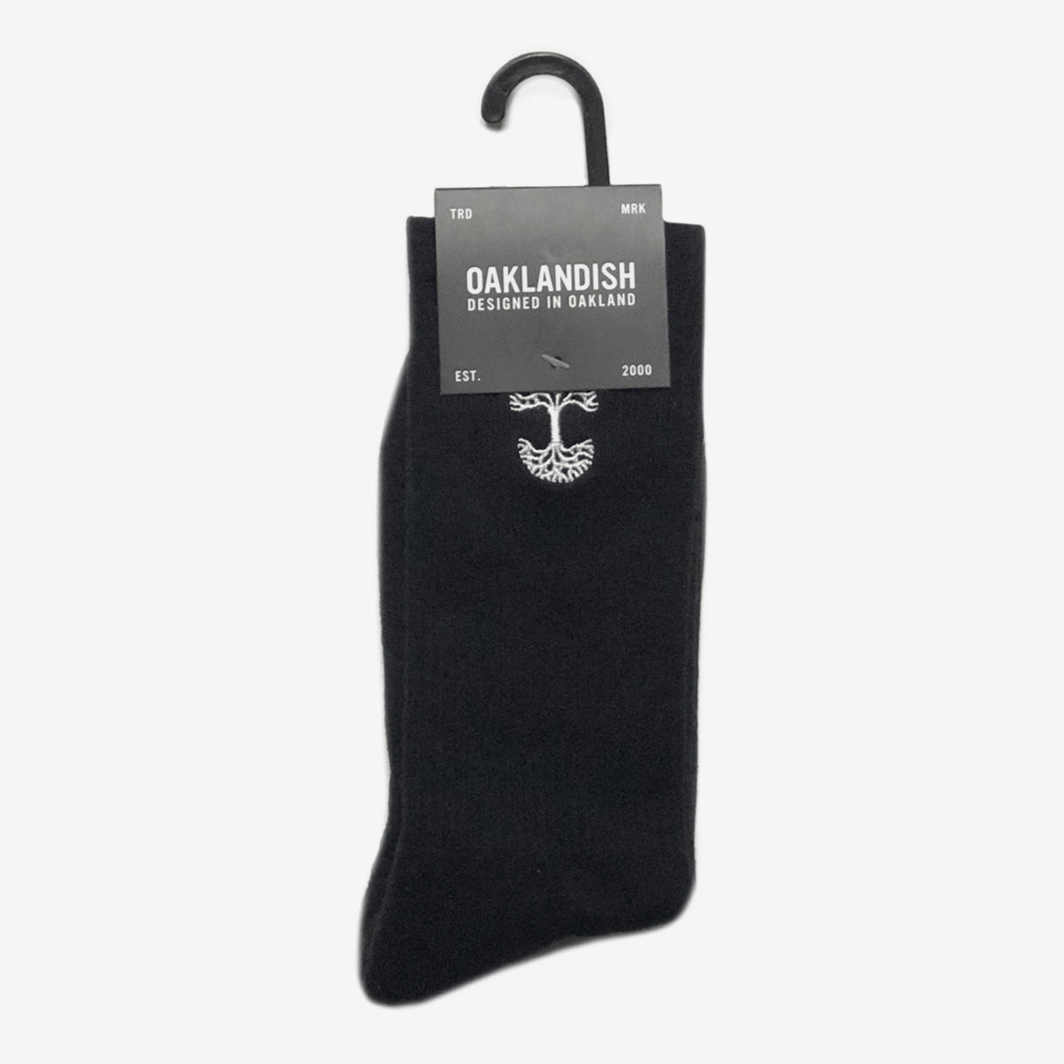 A single black Embroidered Logo Sock from Oaklandish is displayed on a hanger, with a label attached at the top. The label reads "OAKLANDISH DESIGNED IN OAKLAND" along with "TRD MRK" on either side. The year "EST. 2000" is also mentioned. An emblem resembling a tree appears on the adult-sized Oaklandish sock below the label.