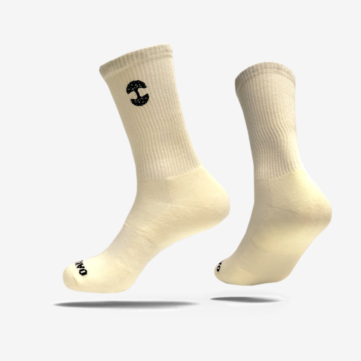 Two off-white cotton crew socks with black logos are suspended mid-air against a white background. The Oaklandish Embroidered Logo Socks are ribbed on the calf area, with one displaying an embroidered stylized "C" near the top and the other sporting a small circular logo on the toe. The overall design is minimalistic and clean.