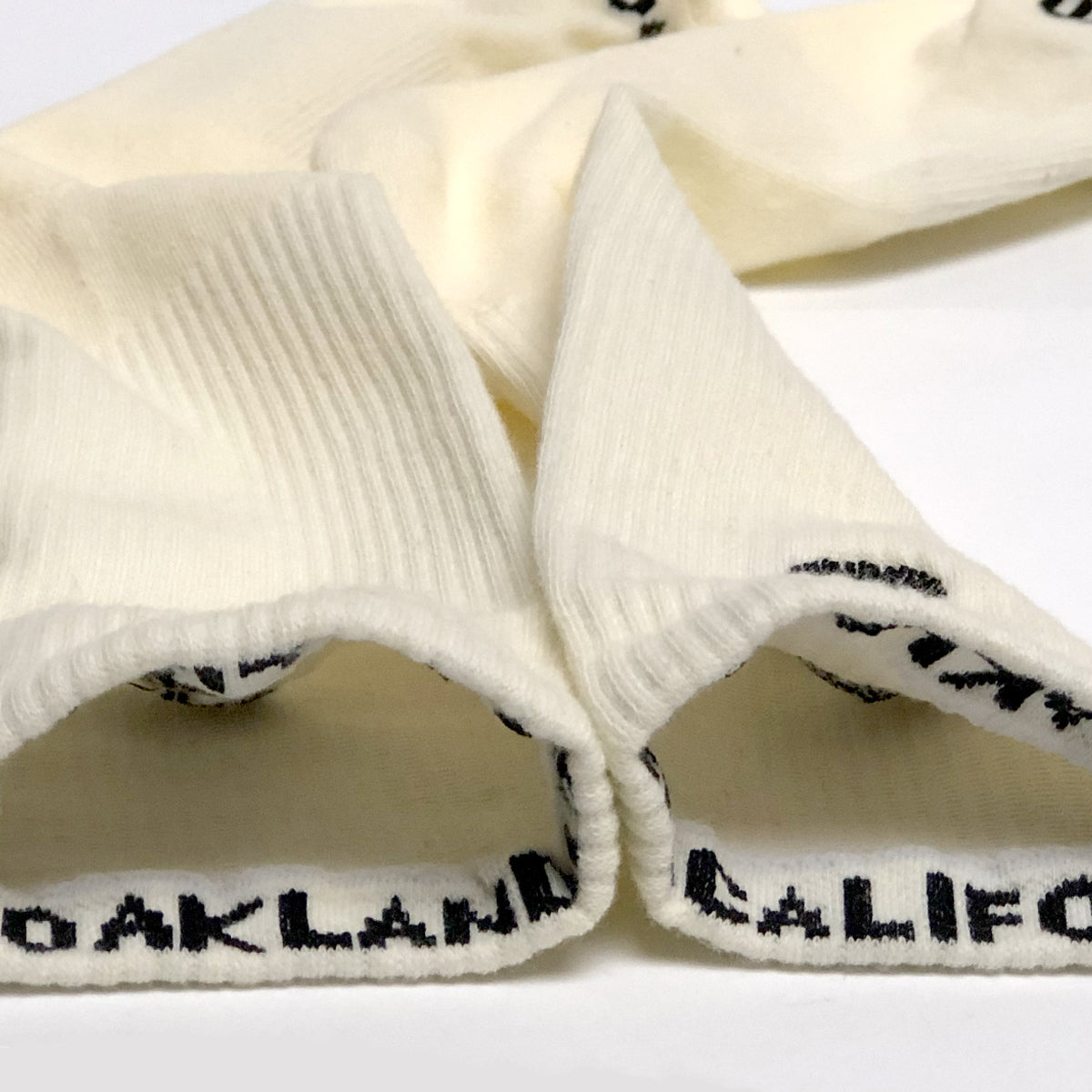 Two off-white cotton crew socks from Oaklandish, known as Embroidered Logo Socks, are laid out with their cuffs turned inside out. The cuffs have a ribbed texture and feature the text "OAKLAND" in black capital letters at the interior edge. These socks are positioned so that the text is partially visible.