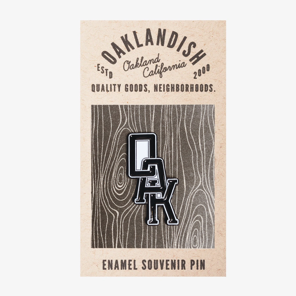 A rectangular card featuring a wood grain pattern in the background holds the Enamel Pin - OAK Monogram from Oaklandish, showcasing "OAK" in black and white letters. Above the pin, text reads: "OAKLANDISH, Estd Oakland California 2000, Quality Goods, Neighborhoods." The card serves as an attractive display for this unique accessory.