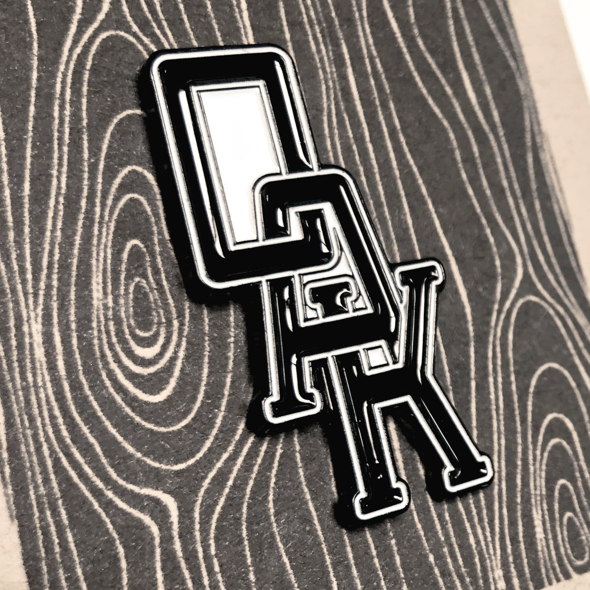 Close up of black OAK wordmark with white trim on enamel lapel pin.