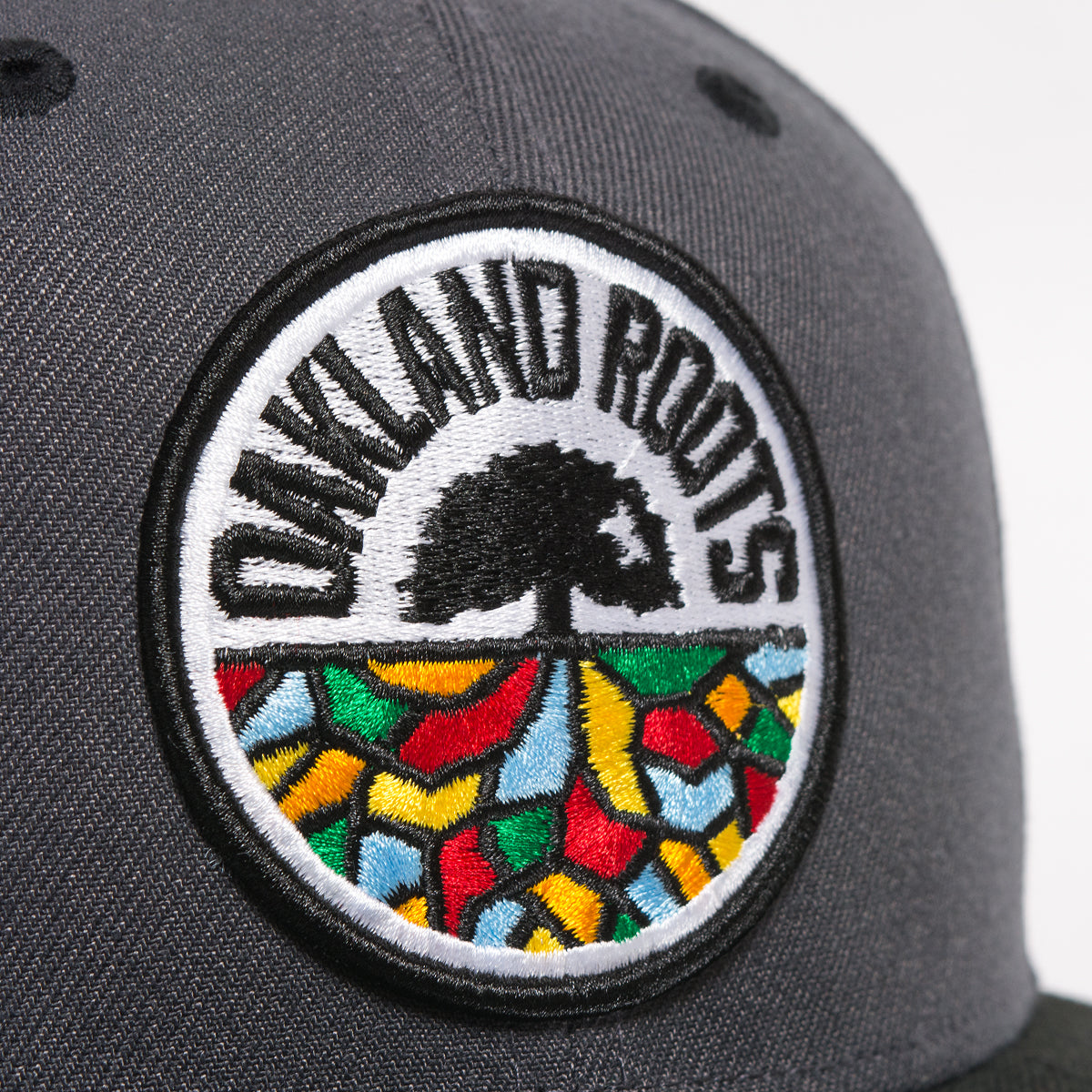 A close-up view of the New Era Oakland Roots SC 59FIFTY Fitted cap in gray features a circular patch with the text "OAKLAND ROOTS" encircling an image of a tree with its roots extending downward. Beneath the roots is a mosaic-like design in red, yellow, green, and blue colors—perfect for any Oakland Roots SC fan.