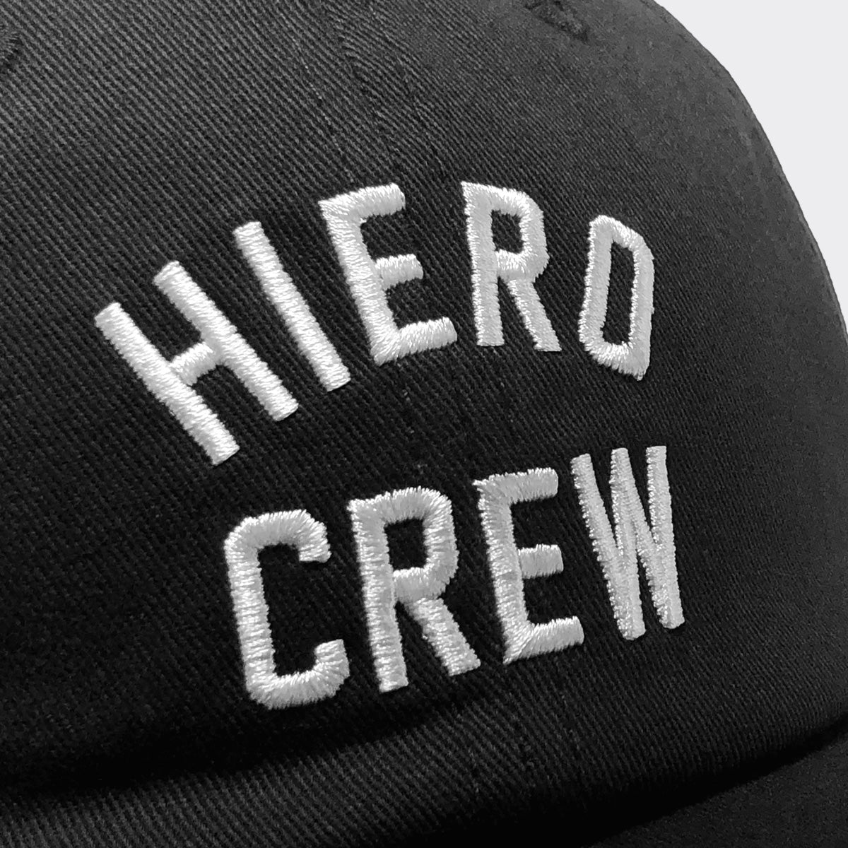 A close-up image features the Hiero Crew Dad Hat by Hieroglyphics, showcasing the text "HIERO CREW" embroidered in white thread. The bold letters stand out against the dark fabric, accentuating the contrast. The plain background ensures all attention is on the detailed stitching and adjustable strapback design of this black dad cap.