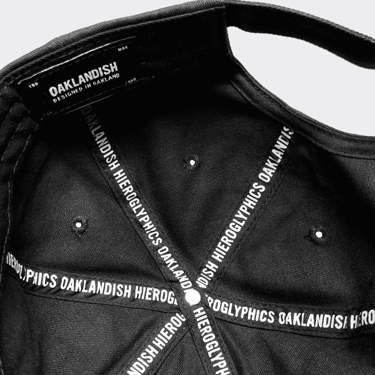 The image shows the interior of the Hiero Crew Dad Hat by Hieroglyphics. The inside is lined with black fabric featuring white text that reads "OAKLANDISH" and "HIEROGLYPHICS." A small label near the front reads "OAKLANDISH DESIGNED IN OAKLAND." The adjustable strapback cap has stitched sections creating a radial pattern.