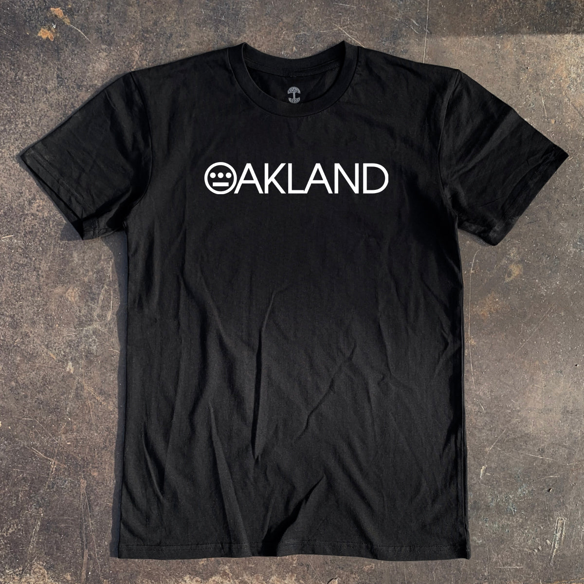 The Hieroakland Tee by Hieroglyphics, a black, 100% cotton T-shirt with a classic fit, lies on a rustic, worn surface. The shirt features "OAKLAND" in white capital letters across the chest with the "O" stylized as a smiley-face graphic, creating a unique design element perfect for Town business.