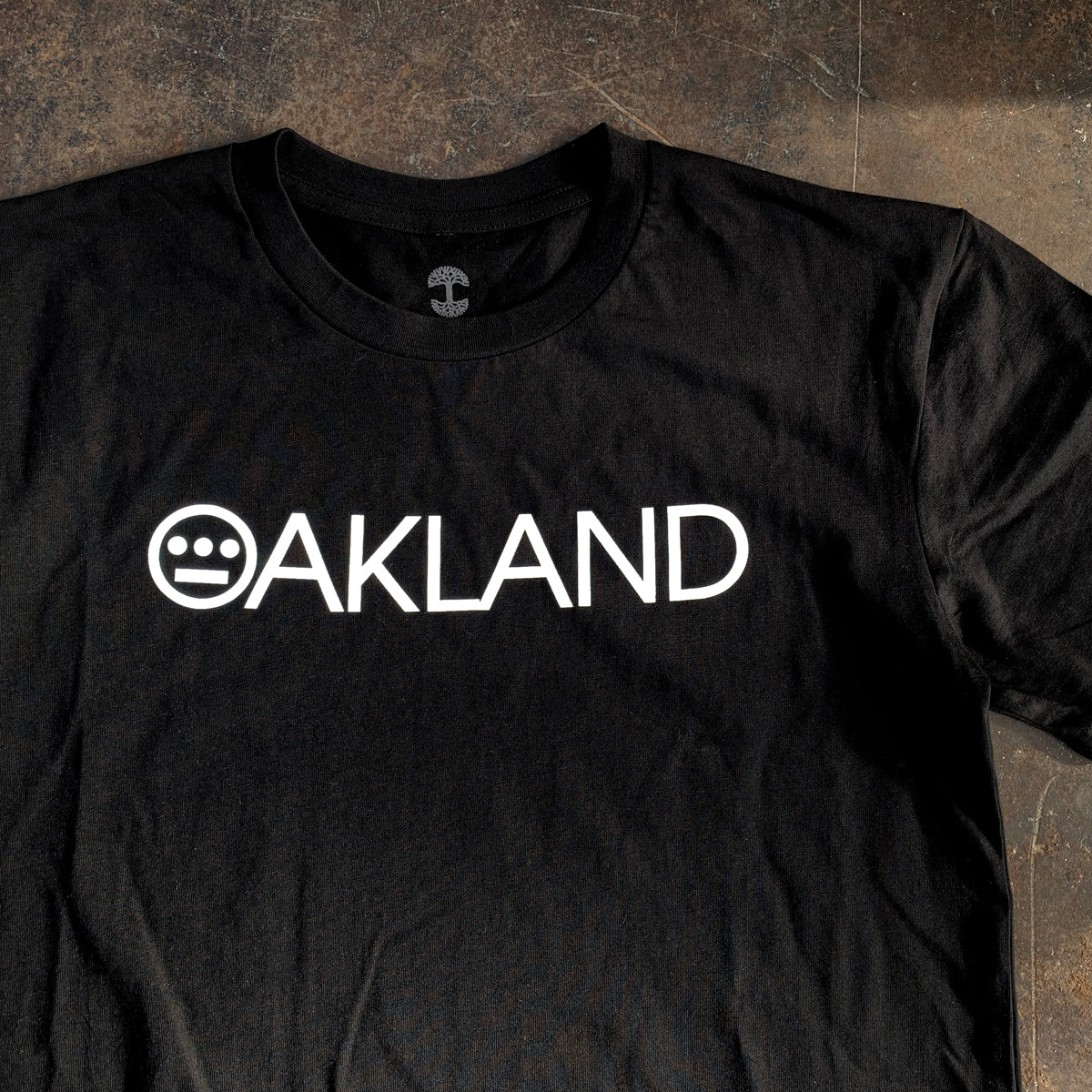 Introducing the Hieroakland Tee from Hieroglyphics—a black, 100% cotton t-shirt featuring "OAKLAND" printed in bold white capital letters across the chest. The letter "O" is uniquely stylized with a circular design incorporating three horizontal lines and two dots, reminiscent of a face. This classic fit t-shirt is photographed laid flat on a dark, textured surface, perfectly representing Town business.