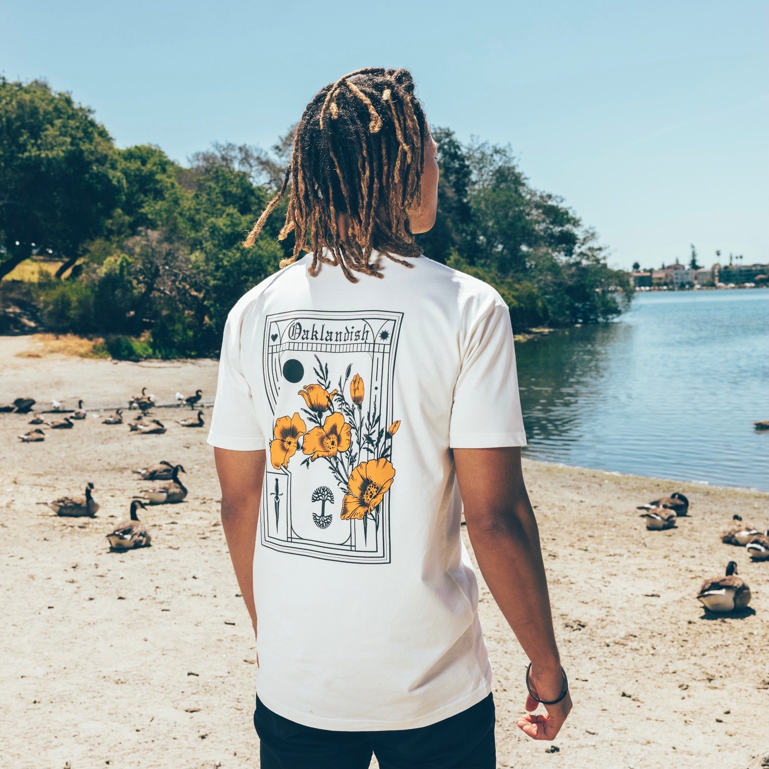 A person with long, braided hair stands on a sandy lakeshore, facing away from the camera. They are wearing an Oaklandish Blossom Tee made of 100% cotton. Ducks are seen on the shore and in the water, with trees and other greenery around the lake under a clear sky.
