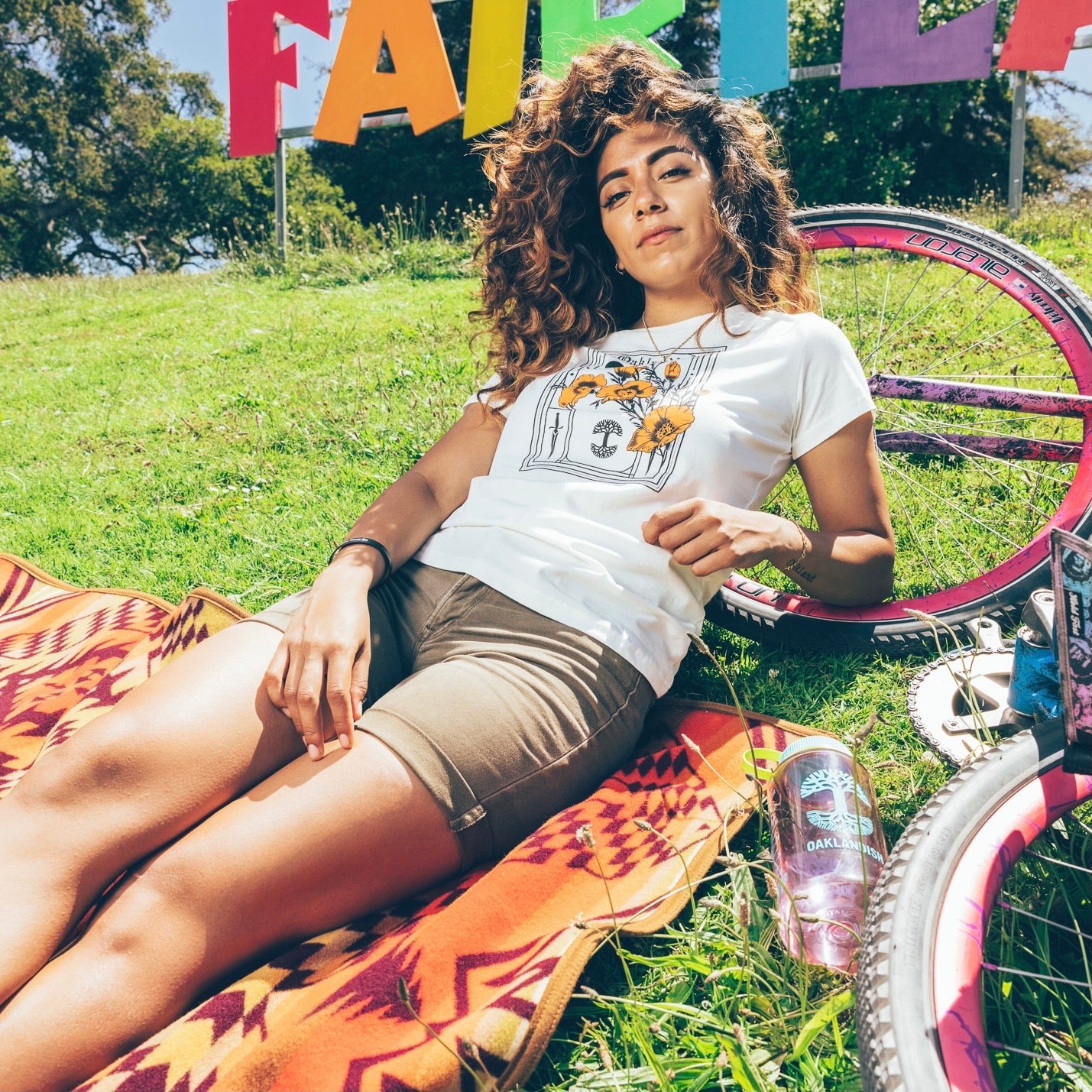 A woman with wavy hair lounges on a patterned blanket on the grass, wearing an Oaklandish Women's Blossom Tee and shorts. Beside her are a bicycle with pink tires and a can of Oak beer. In the background, colorful letters spell "FAIR PLAY." Trees are visible behind the sign.