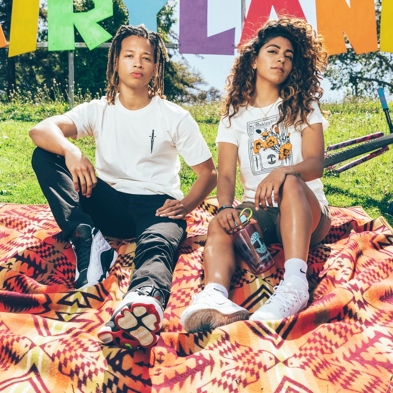 A young man and woman sit on a vibrant patterned blanket on a sunny day, with a colorful sign in the background. The man, dressed in black pants and a classic fit 100% cotton t-shirt, has dreadlocks. The woman, holding a clear bottle, wears shorts and an Oaklandish Blossom Tee that complements her curly hair.