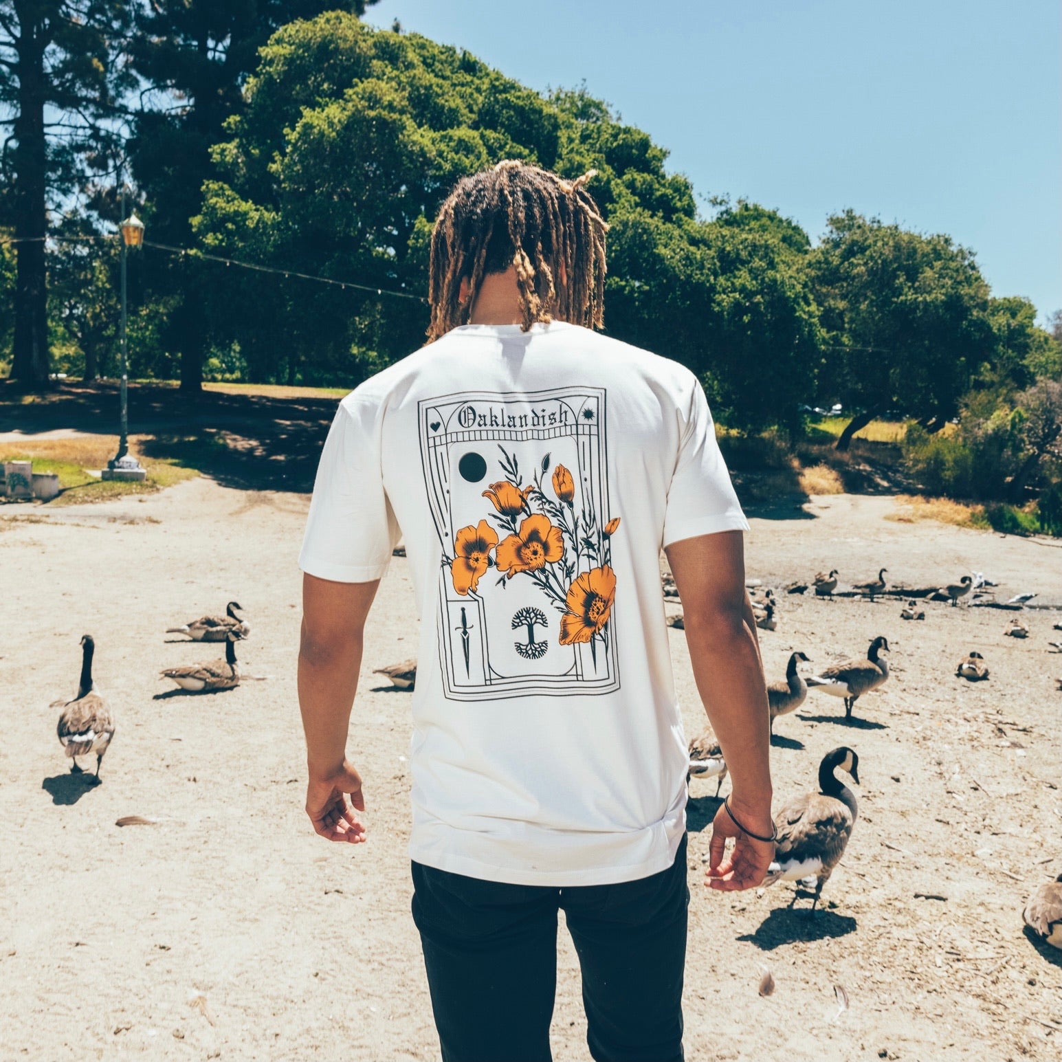 A person with long, light brown hair is standing outdoors with their back facing the camera, wearing the Blossom Tee by Oaklandish. The 100% cotton white t-shirt features a floral graphic of poppies and the word "Outlandish." Surrounded by geese and trees, with a clear sunny sky and a lantern in the background.
