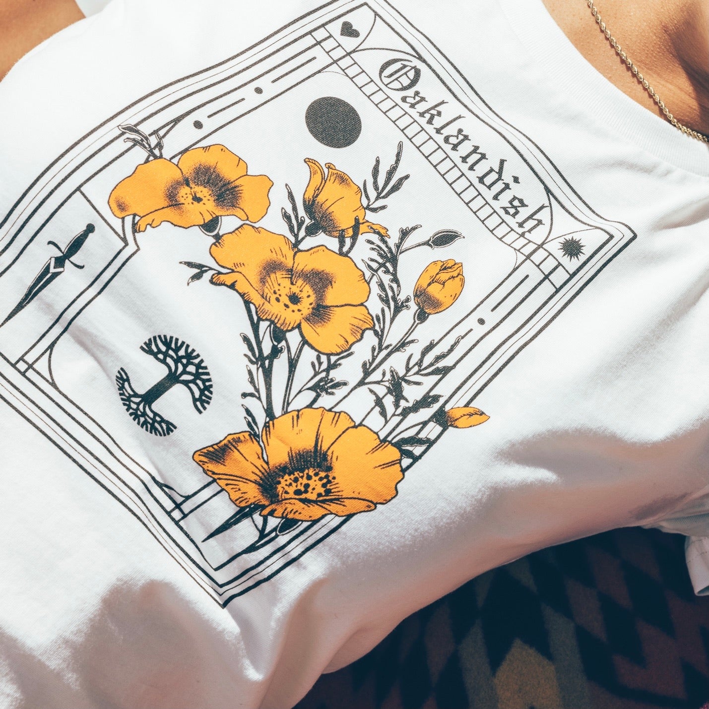 Close-up of a person wearing the Women's Blossom Tee from Oaklandish, a 100% cotton white t-shirt featuring an intricate design with yellow-orange poppies and botanical elements. The classic fit design resembles a vintage botanical print, with the word "Outlandish" at the top, bordered by decorative lines and small icons, including a crescent moon.