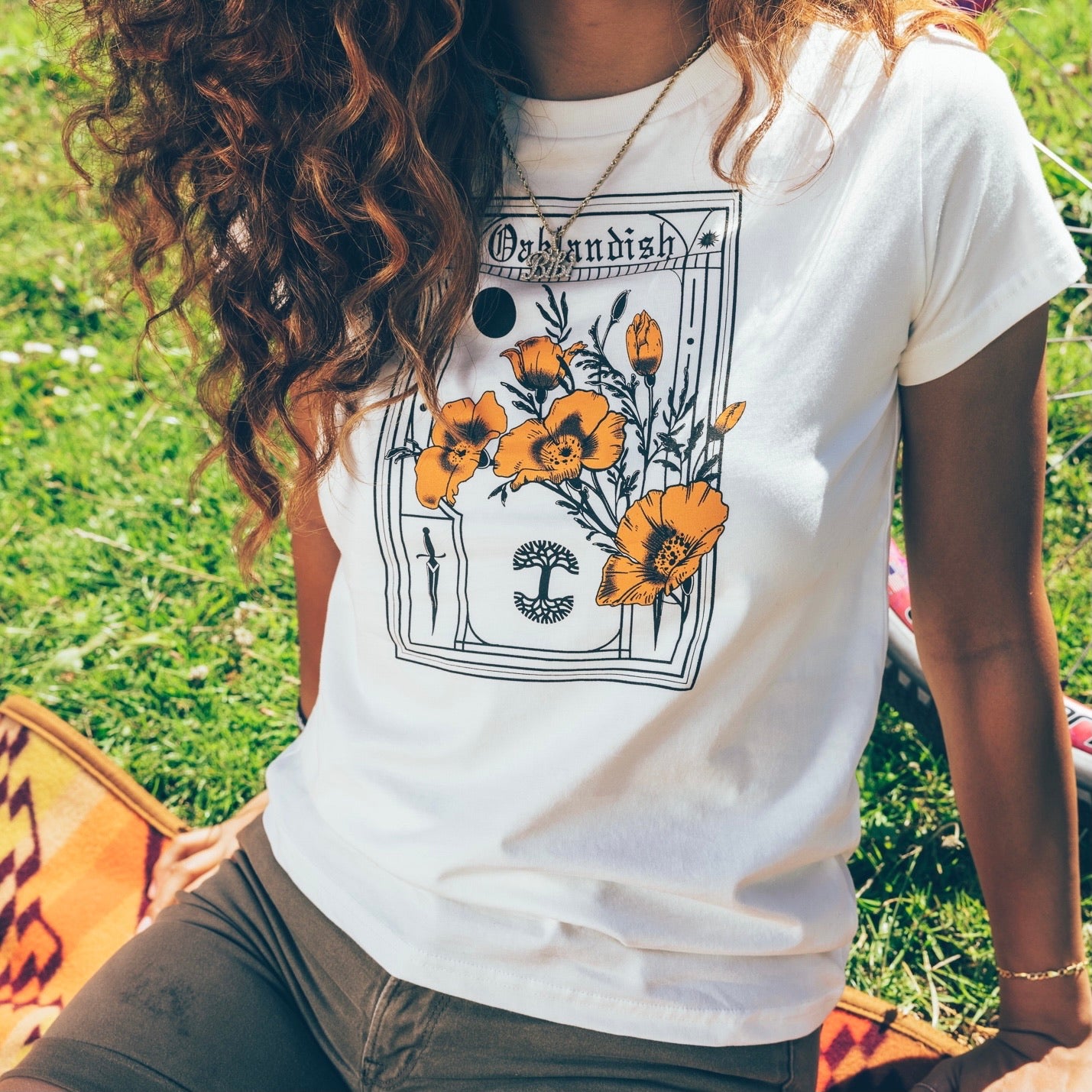 A person with long, wavy hair sits on the grass, wearing an intricately designed Women's Blossom Tee from Oaklandish that features vibrant orange flowers and the word "Outlandish." They pair it with olive green pants, while a colorful blanket is partially visible beside them on the ground.