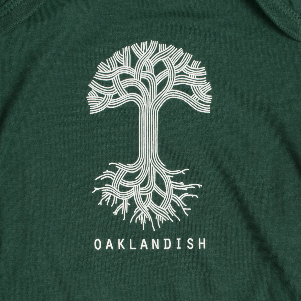 A close-up of the Infant Oaklandish Classic Logo One-Piece, made from soft ring-spun cotton in a dark green color, features a white graphic of an abstract tree with intricate, symmetrical branches and roots. Below the tree design, the word "OAKLANDISH" is printed in capital letters.