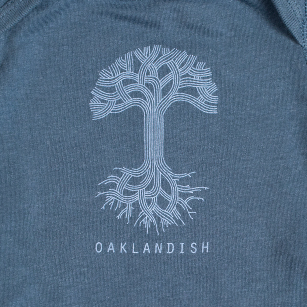The Infant Oaklandish Classic Logo One-Piece from Oaklandish is a blue-gray fabric baby romper featuring an intricate white tree design at its center. The cotton tree showcases intertwining branches and roots, forming a symmetrical pattern. Below the tree graphic, "OAKLANDISH" is printed in all capital letters.