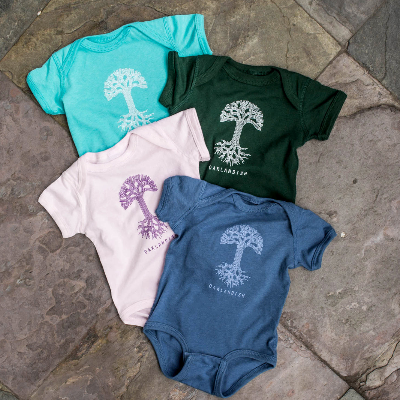 Four "Infant Oaklandish Classic Logo One-Piece" rompers from the Oaklandish brand are displayed on a stone-tiled floor, arranged in a fan shape. The teal romper is in the top left, dark green in the top right, pink at the bottom left, and blue at the bottom right. Each romper features a white tree design with "OAKLANDISH" printed beneath it and boasts a reinforced closure made from ring-spun cotton.