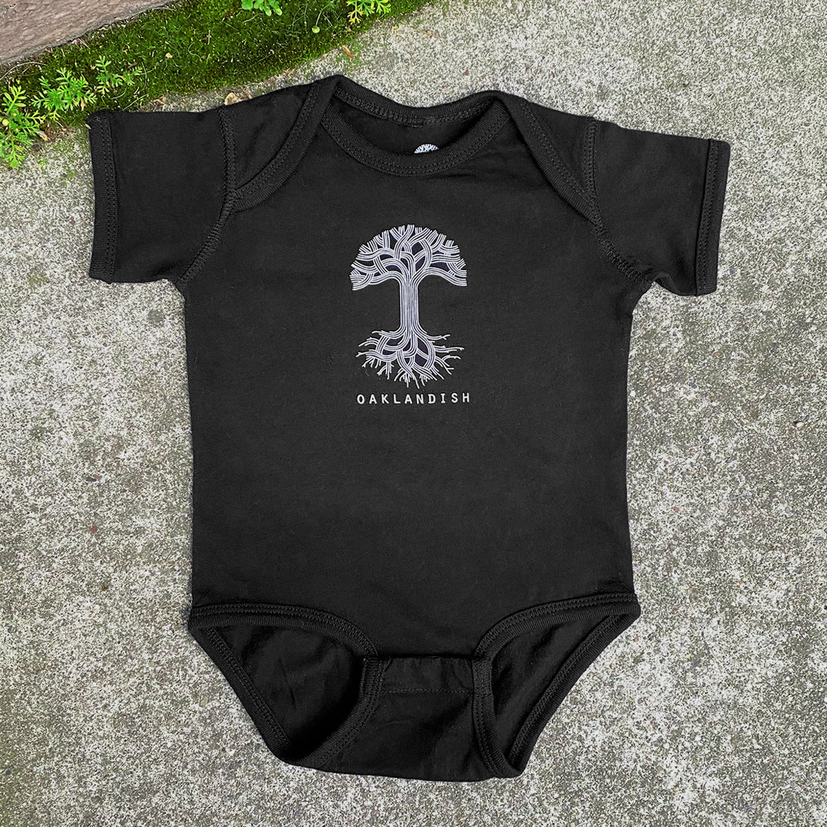 The Infant Oaklandish Classic Logo One-Piece by Oaklandish, a black romper made from 100% cotton with short sleeves, lies flat on a concrete surface. The onesie features a white graphic in the center depicting a tree with its branches and roots spread out. Below the tree graphic is the word "OAKLANDISH" in capital letters. Green foliage can be seen in the top left corner.