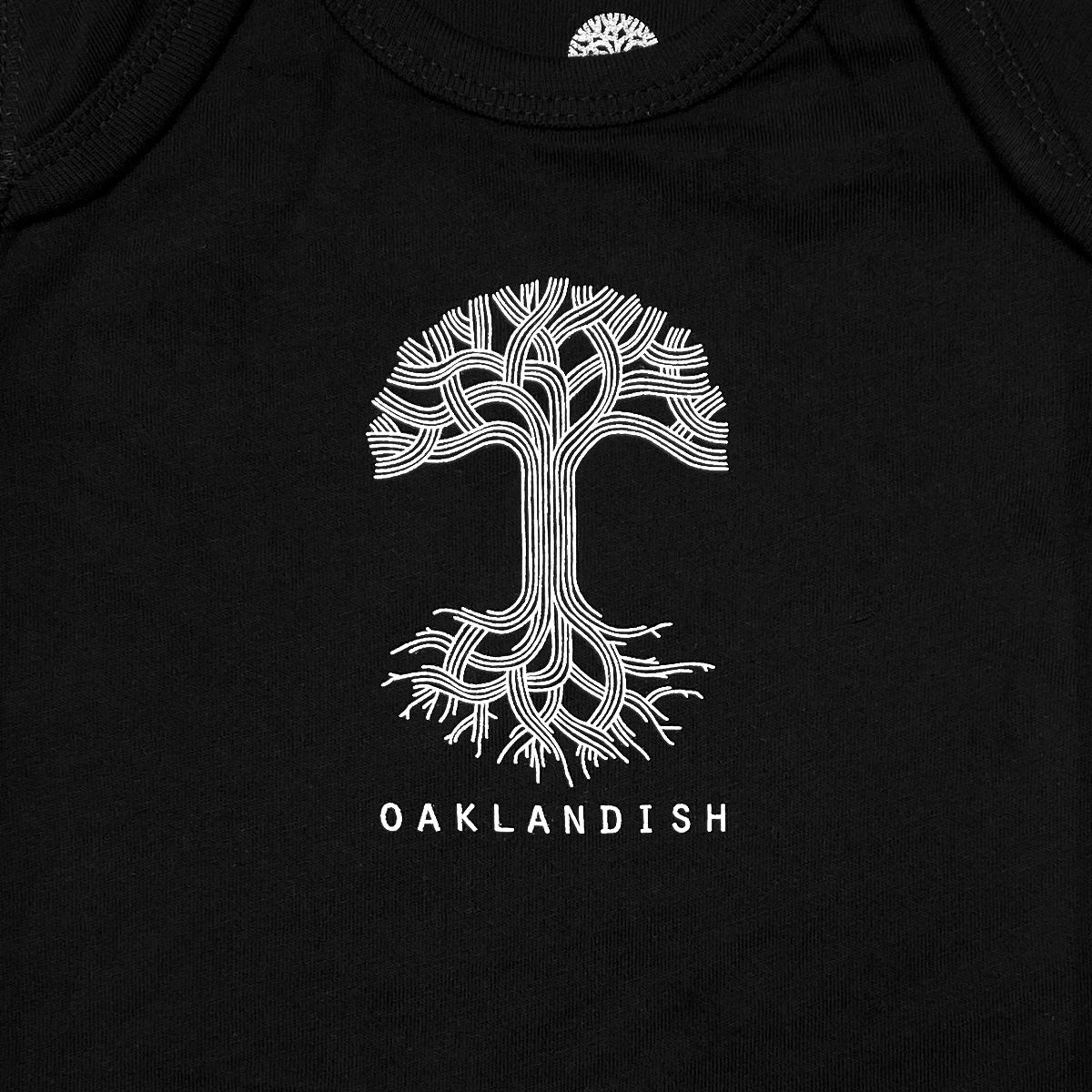The Infant Oaklandish Classic Logo One-Piece by Oaklandish is a black romper made from 100% cotton. It features a detailed white graphic design of a tree with intricate, interwoven branches and roots, along with reinforced snap closures for added durability. Below the tree, "OAKLANDISH" is printed in bold white uppercase letters.